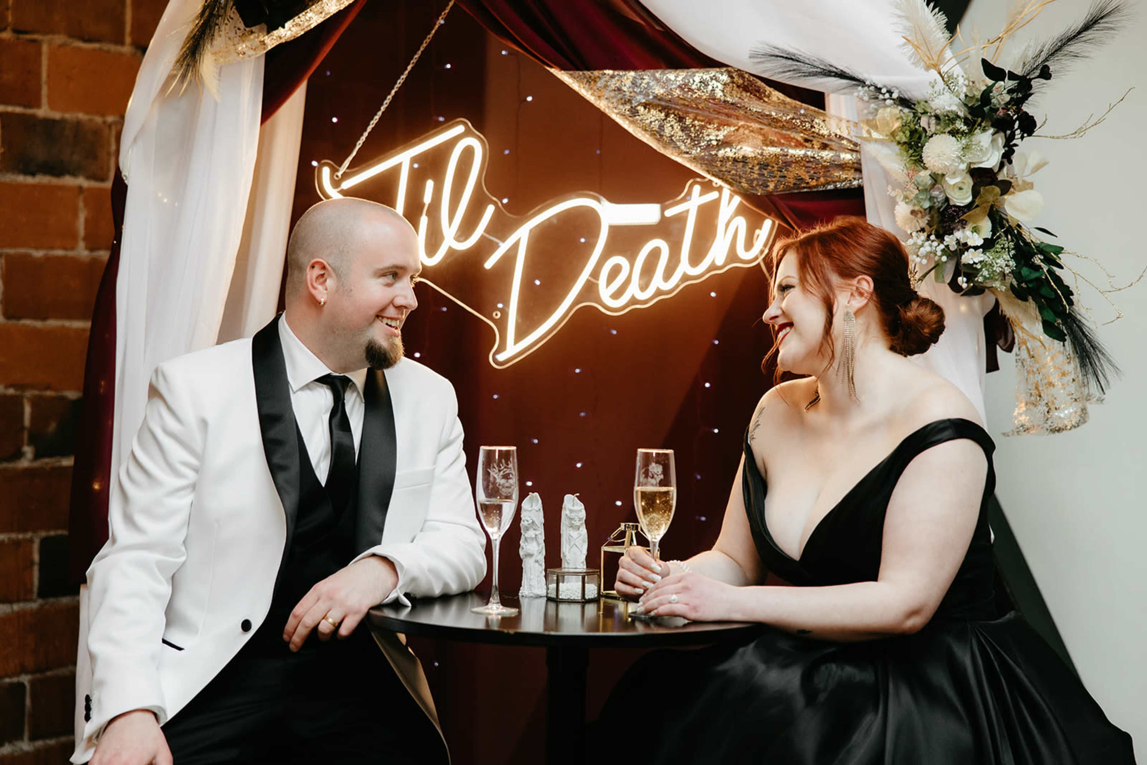 A couple in formal attire sits at a small table with champagne flutes, smiling at each other in front of a "Til Death" neon sign.