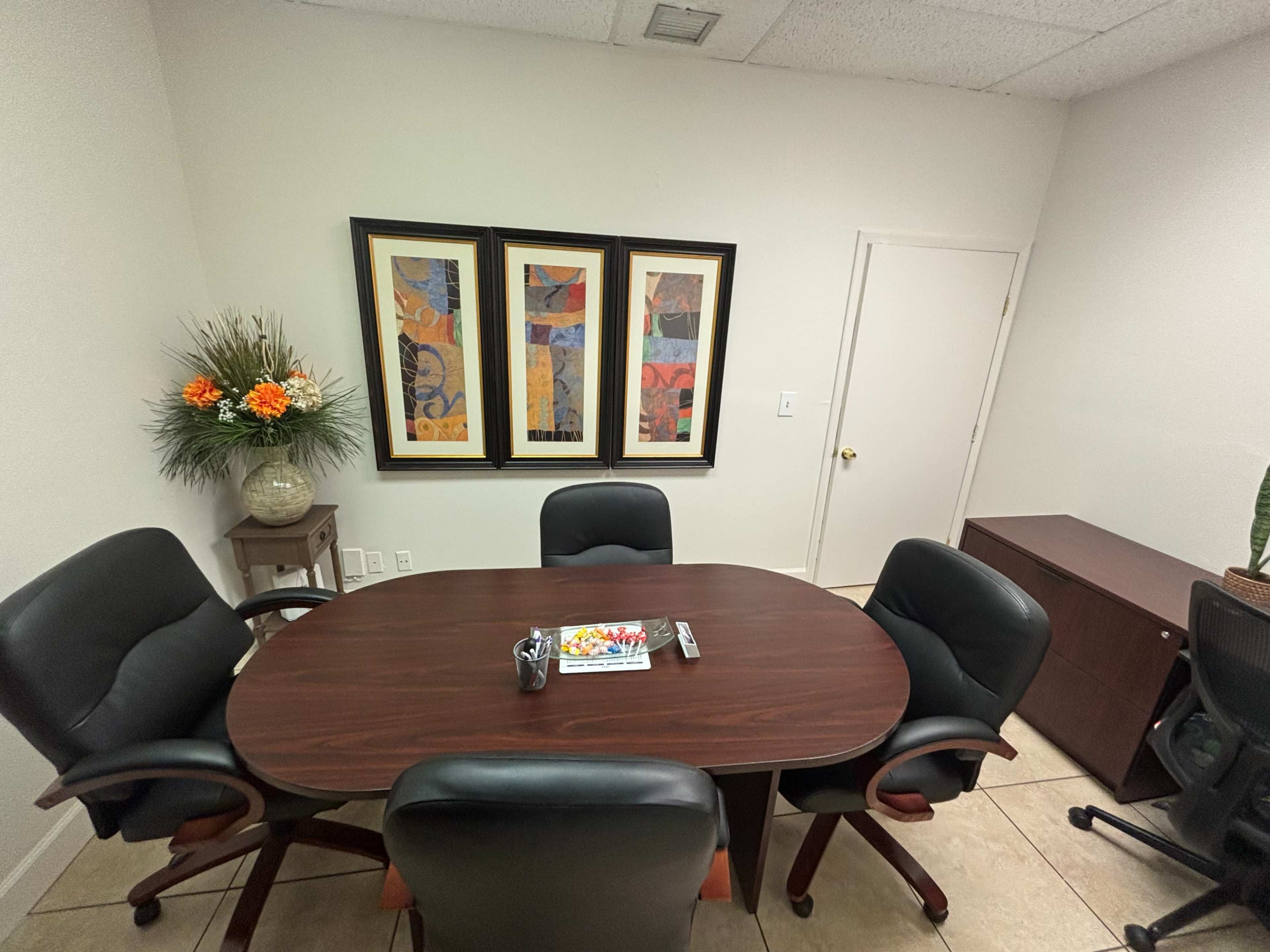 The image shows a small conference room with an oval table surrounded by four black chairs, framed artwork on the wall, and a large plant in the corner.