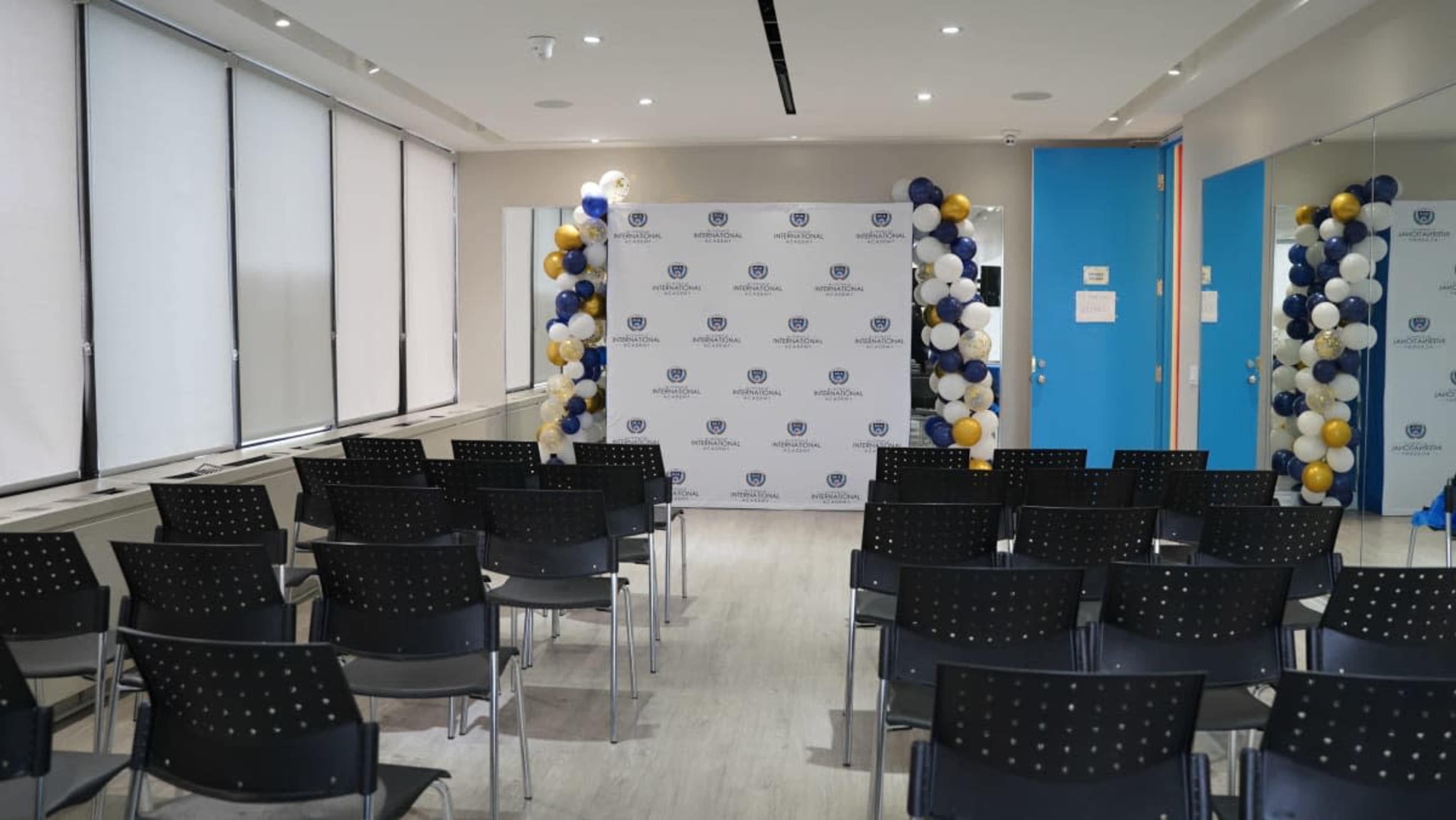 A conference room is set up with rows of black chairs facing a decorated backdrop featuring a logo, flanked by clusters of balloons in blue, gold, and white.