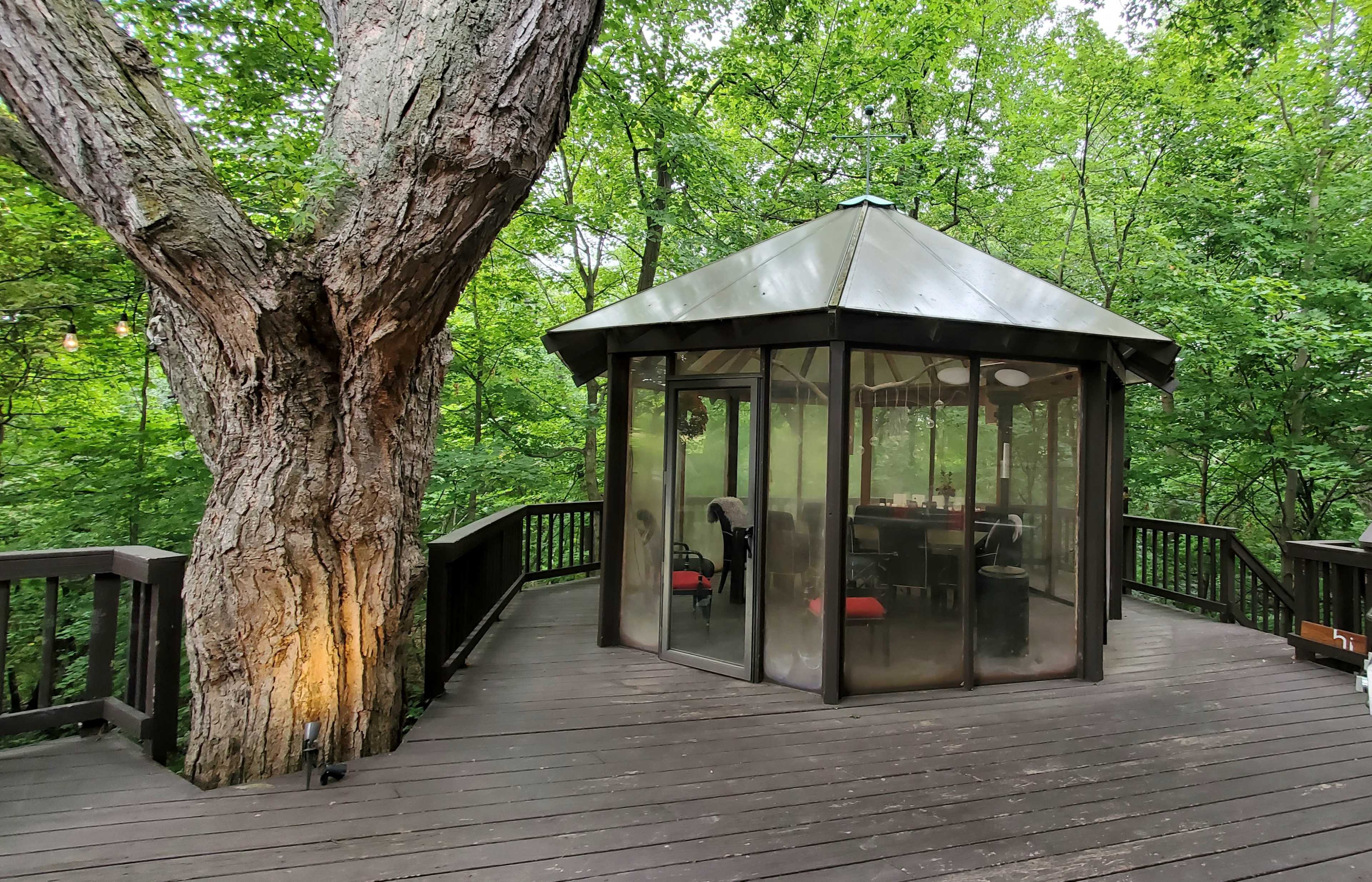 A hexagonal gazebo with a glass enclosure sits on a wooden deck surrounded by lush green trees.