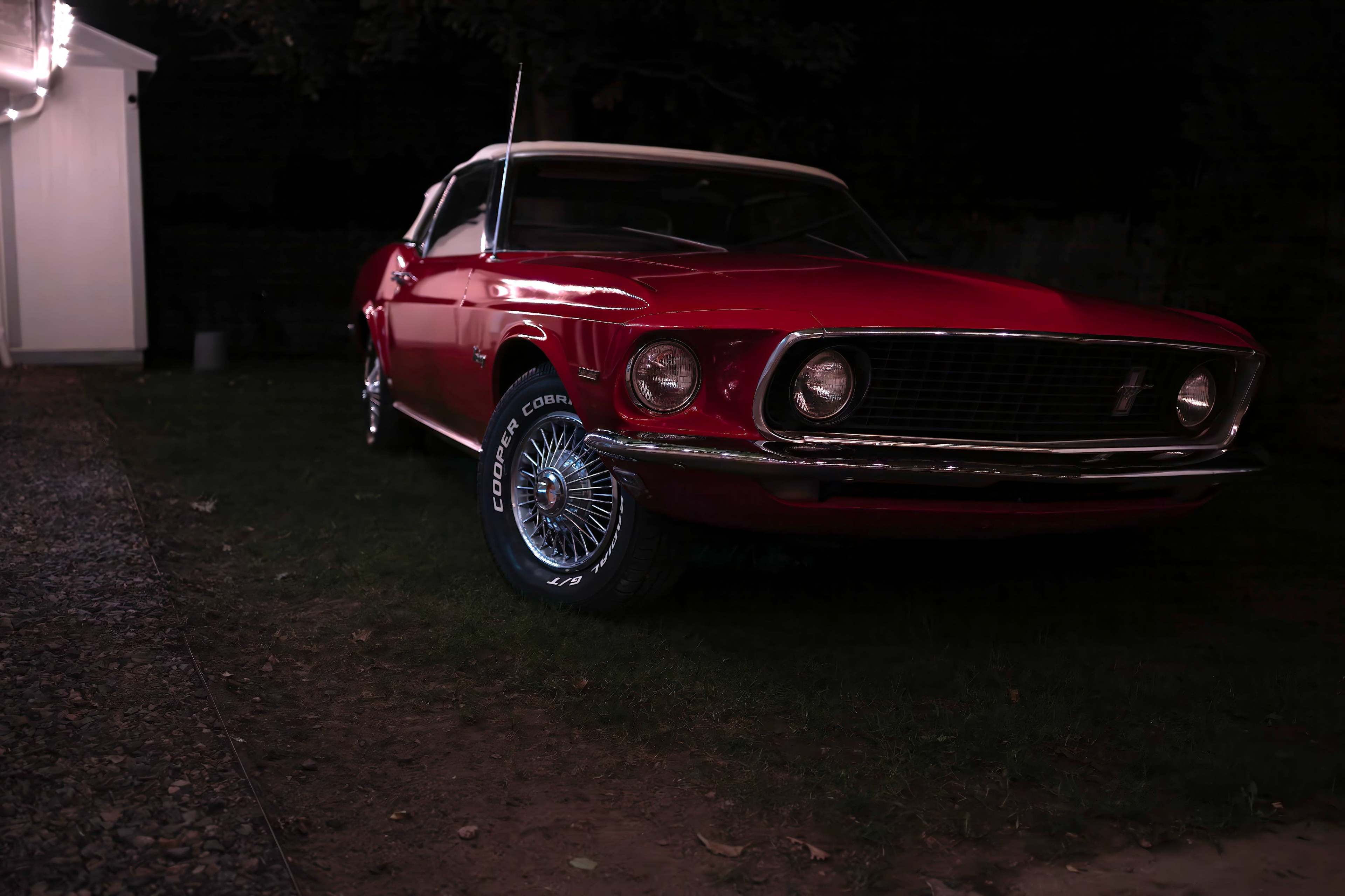 A red 1969 Mustang convertible is parked on a grassy area at night, illuminated by nearby outdoor lights.