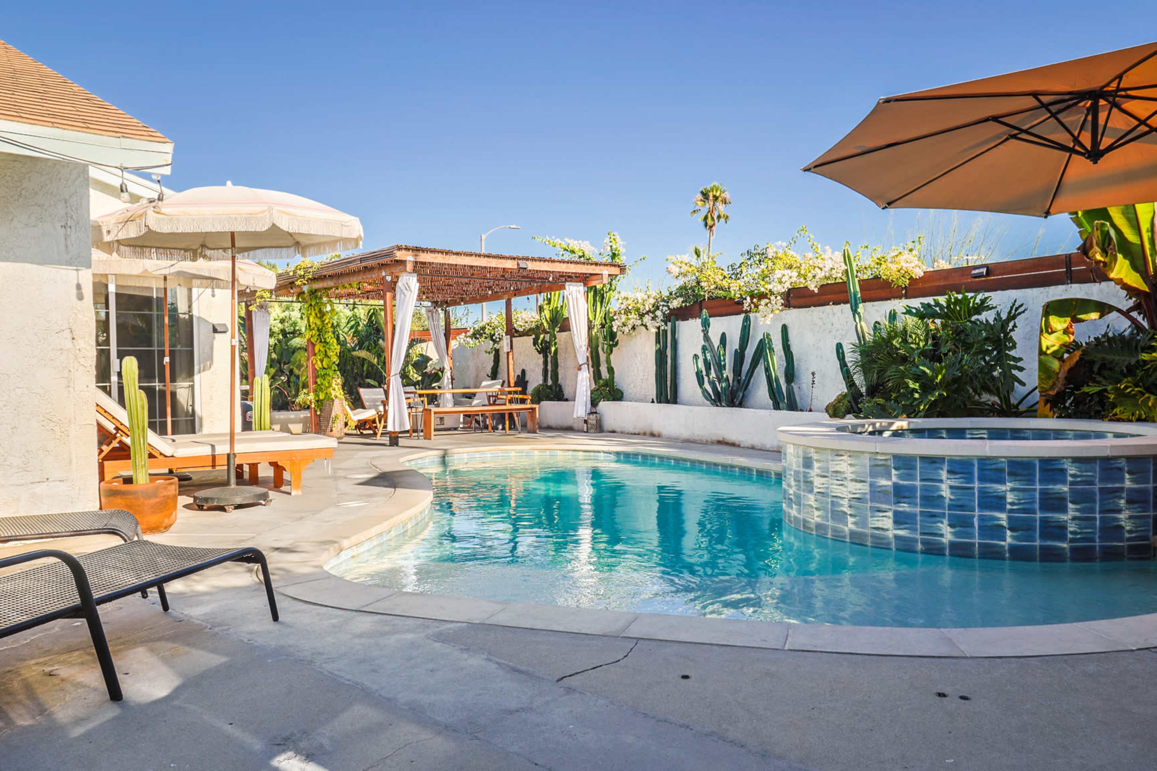 A backyard with a swimming pool, a shaded seating area with a pergola, and various plants surrounding the space.