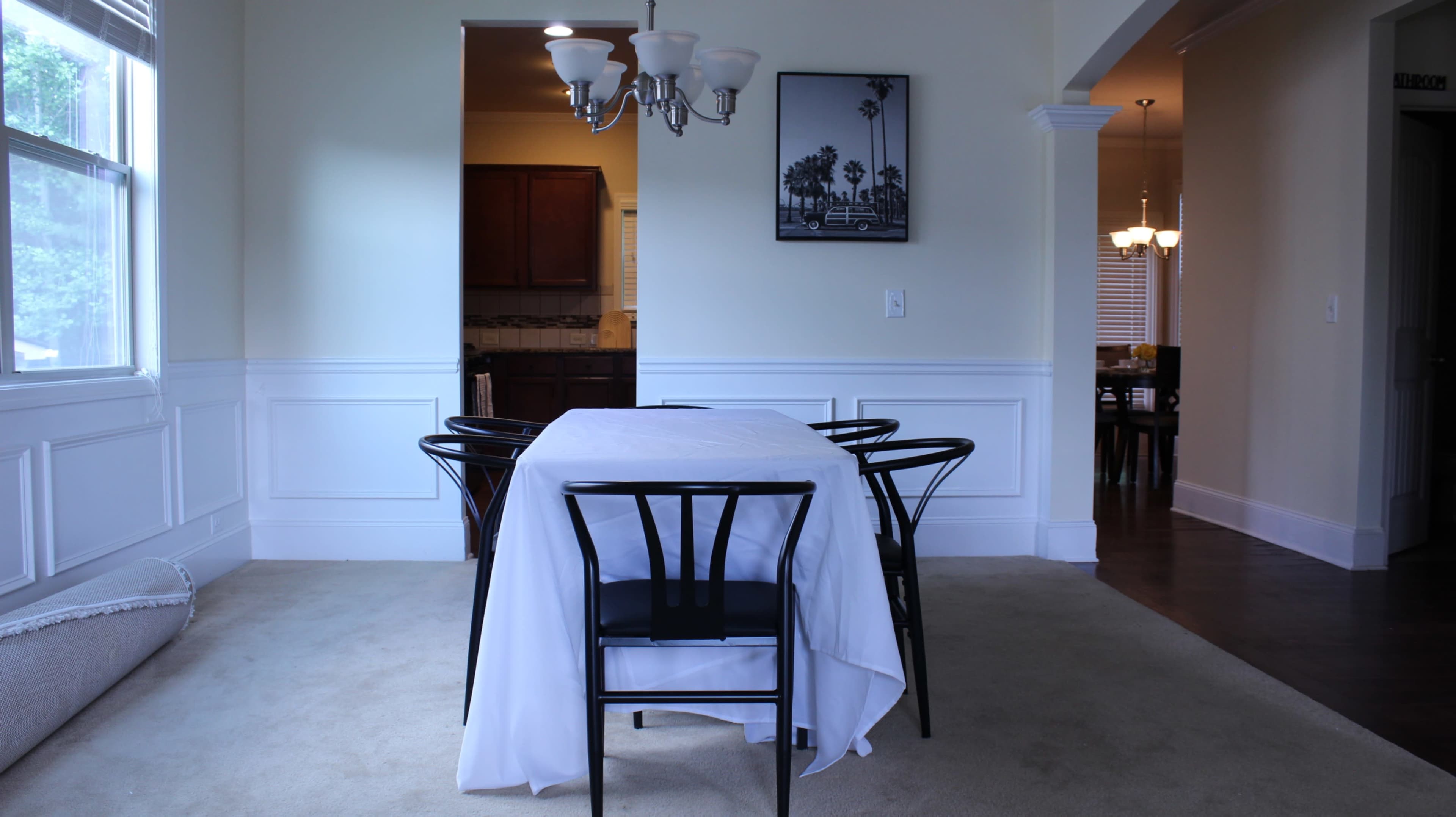 The image shows a dining area with a white tablecloth on a table surrounded by black chairs, leading into a kitchen area through an open archway.