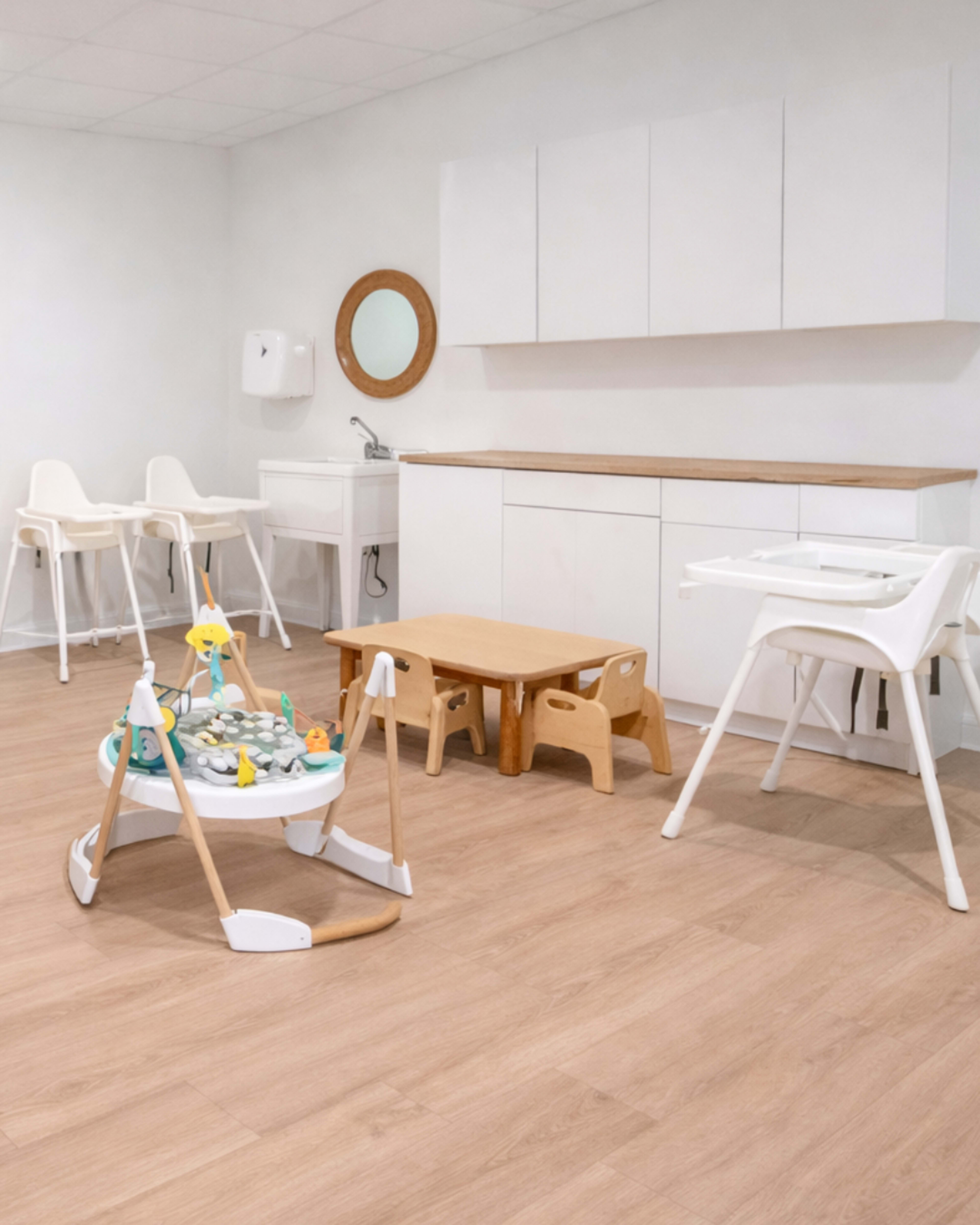 The image shows a bright, minimally furnished room designed for children, featuring a wooden floor, a small table, high chairs, and a playpen with toys.