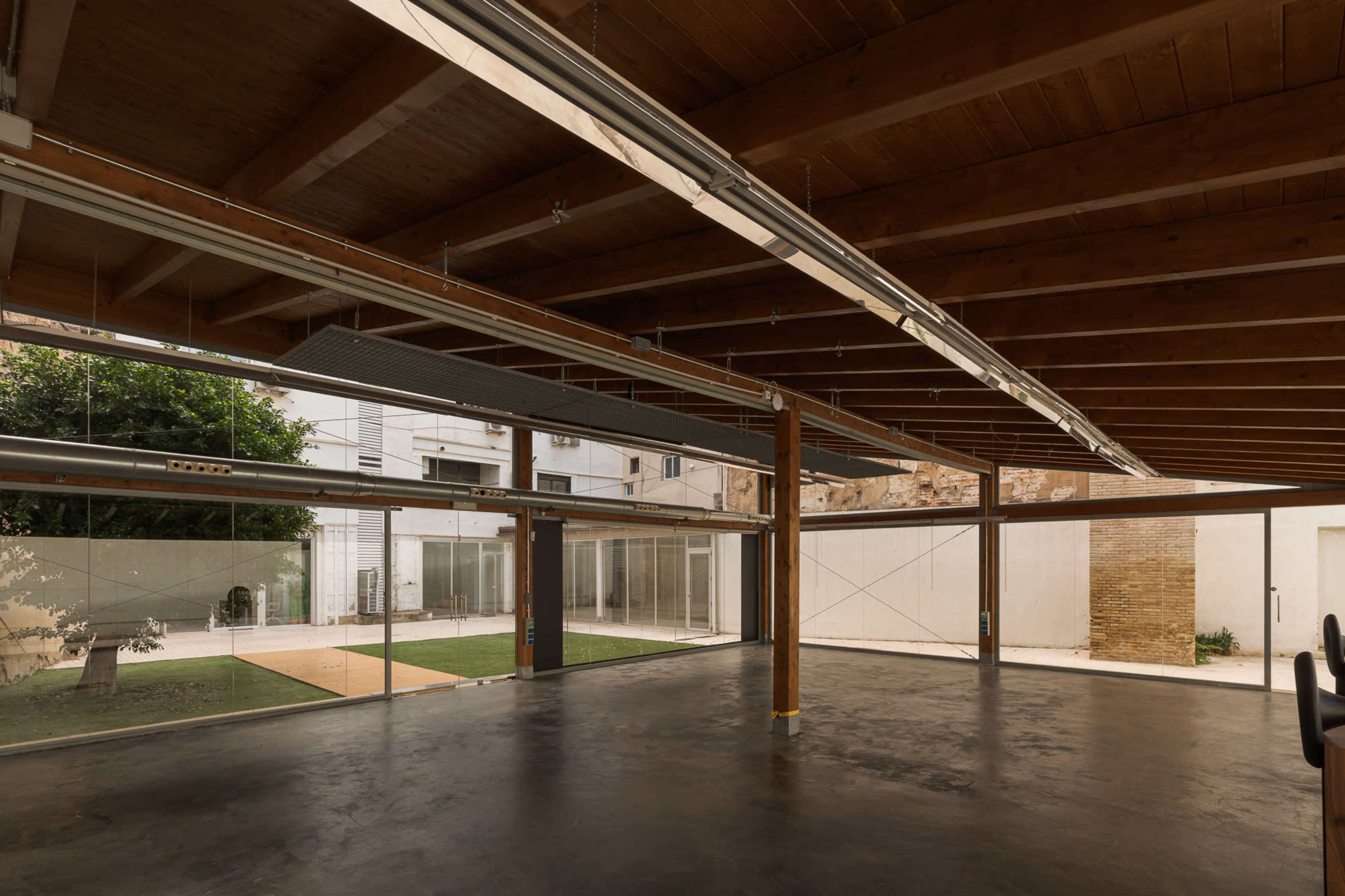 The image shows an open interior space with wooden beams, large glass panels, and a view of a grass area outside.