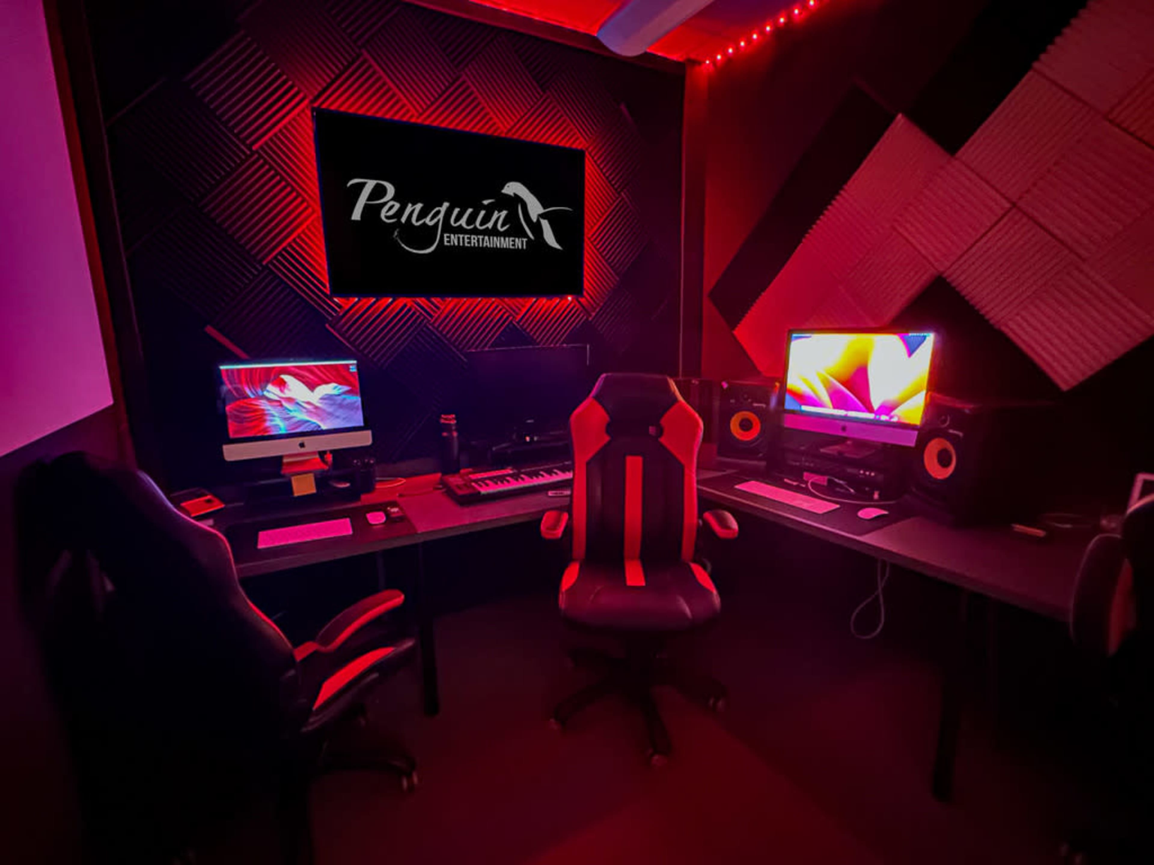 A music production studio featuring two computer workstations, audio speakers, and a prominently displayed logo for "Penguin Entertainment" against a dark background with red lighting.