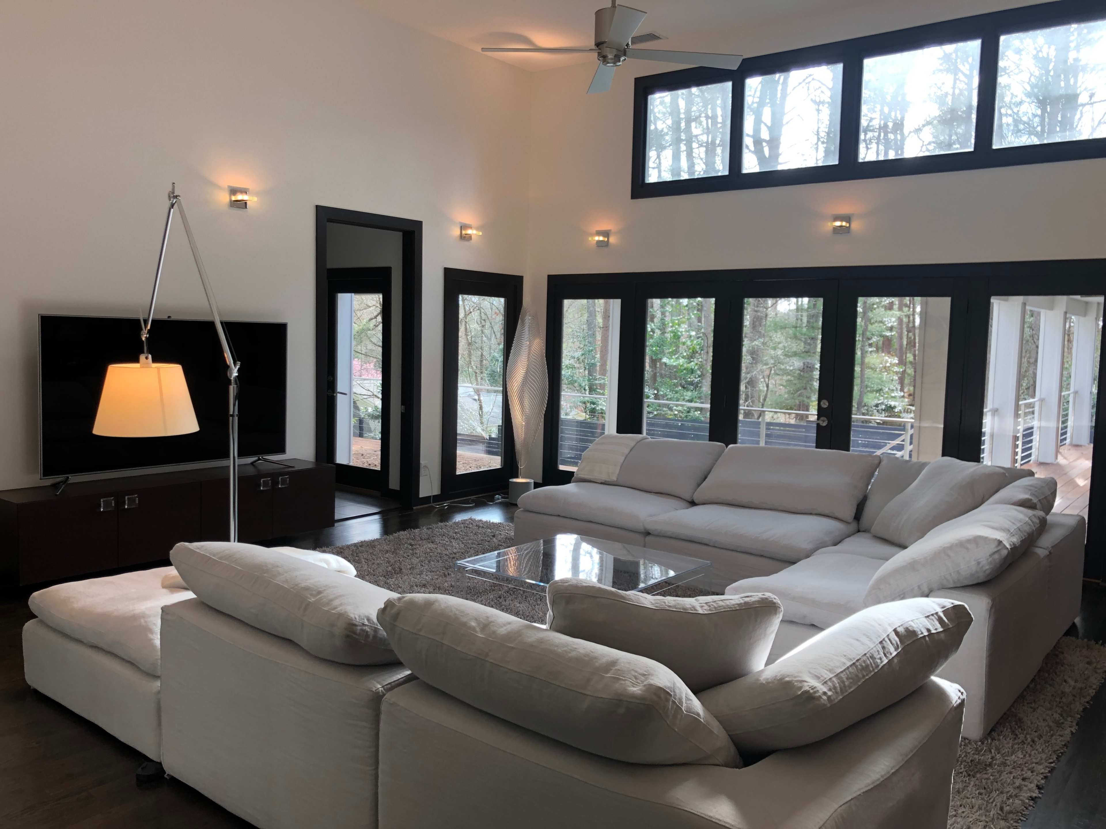 The image shows a modern living room with a large sectional sofa, a coffee table, and floor-to-ceiling windows overlooking a forested area.