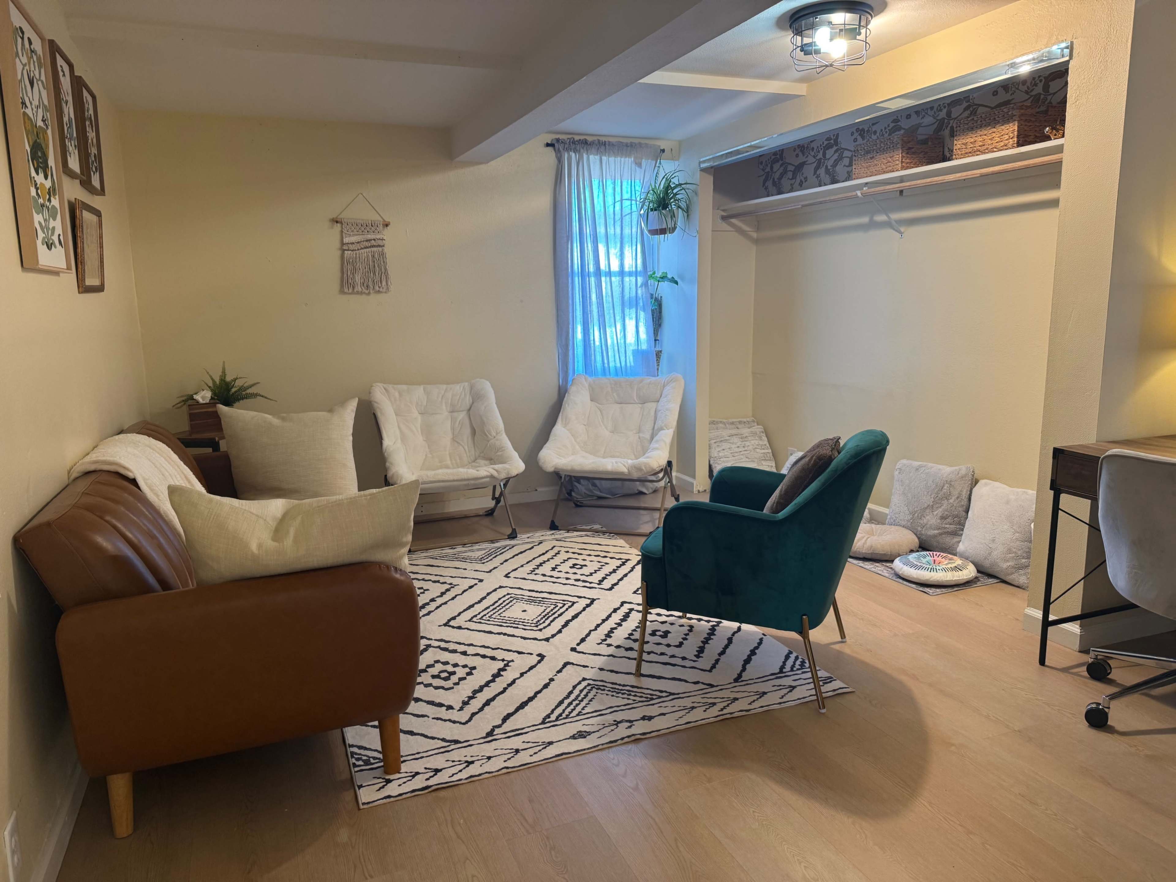The image shows a cozy room with two white chairs and a green armchair arranged around a patterned rug, alongside a brown couch and minimal decorative elements.