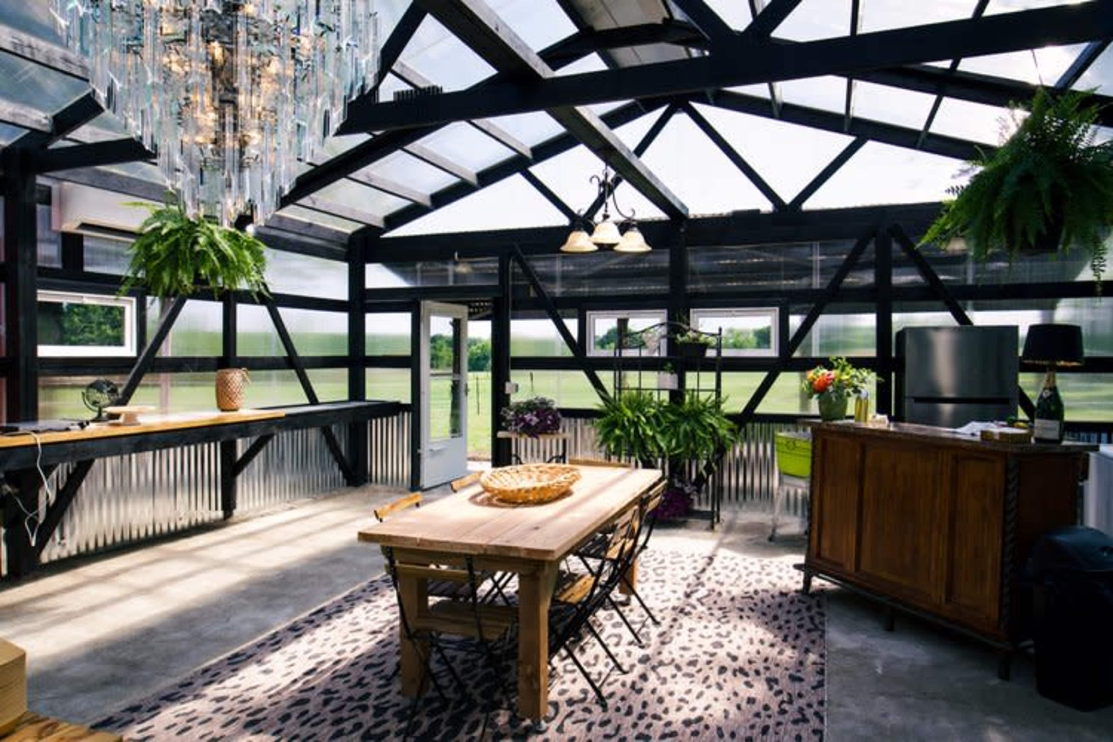 A spacious greenhouse with a wooden dining table, surrounded by plants and natural light streaming through large windows.