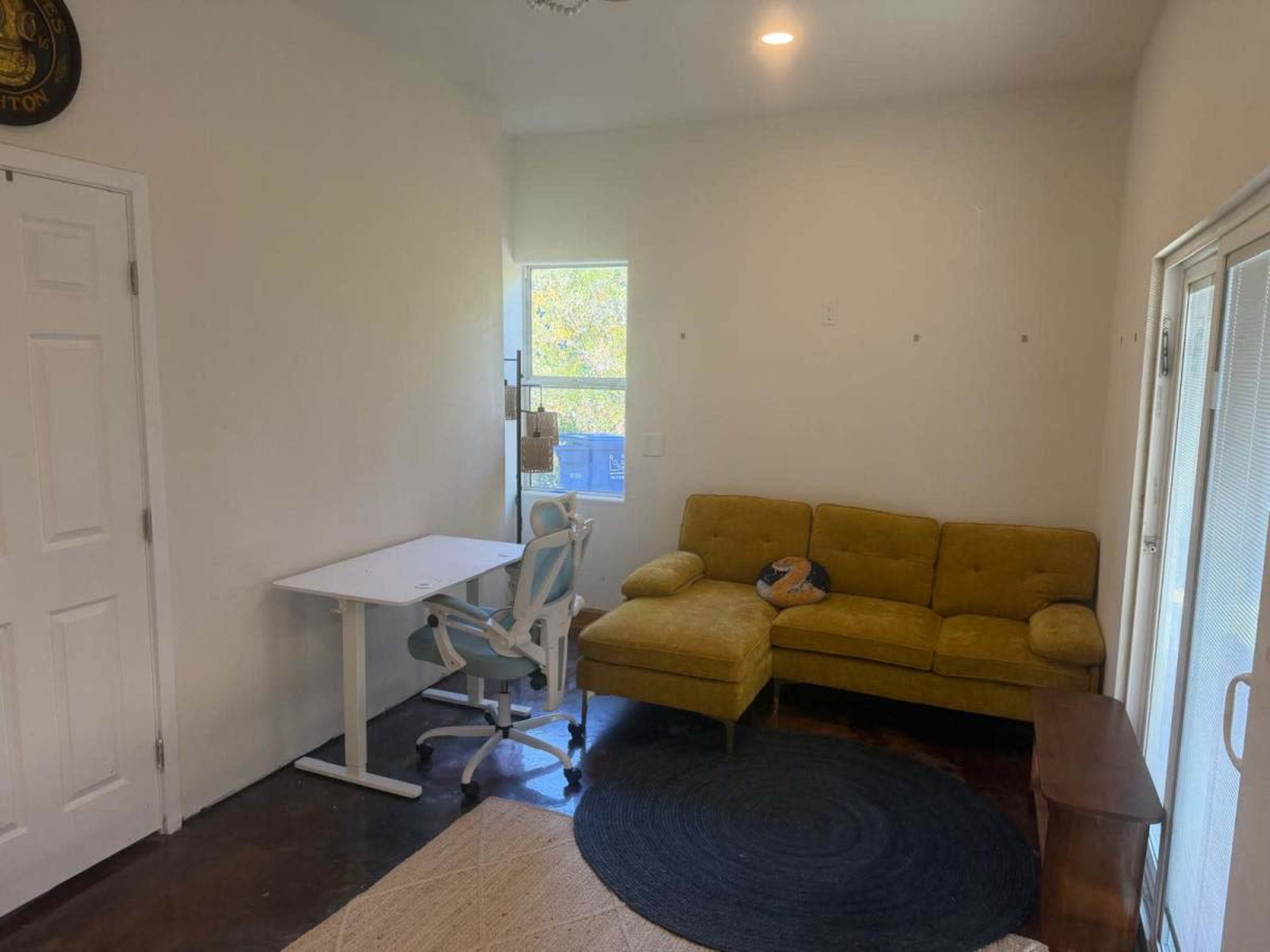 The image shows a small, sparsely furnished room with a yellow sectional couch, a desk with a chair, and a round rug.