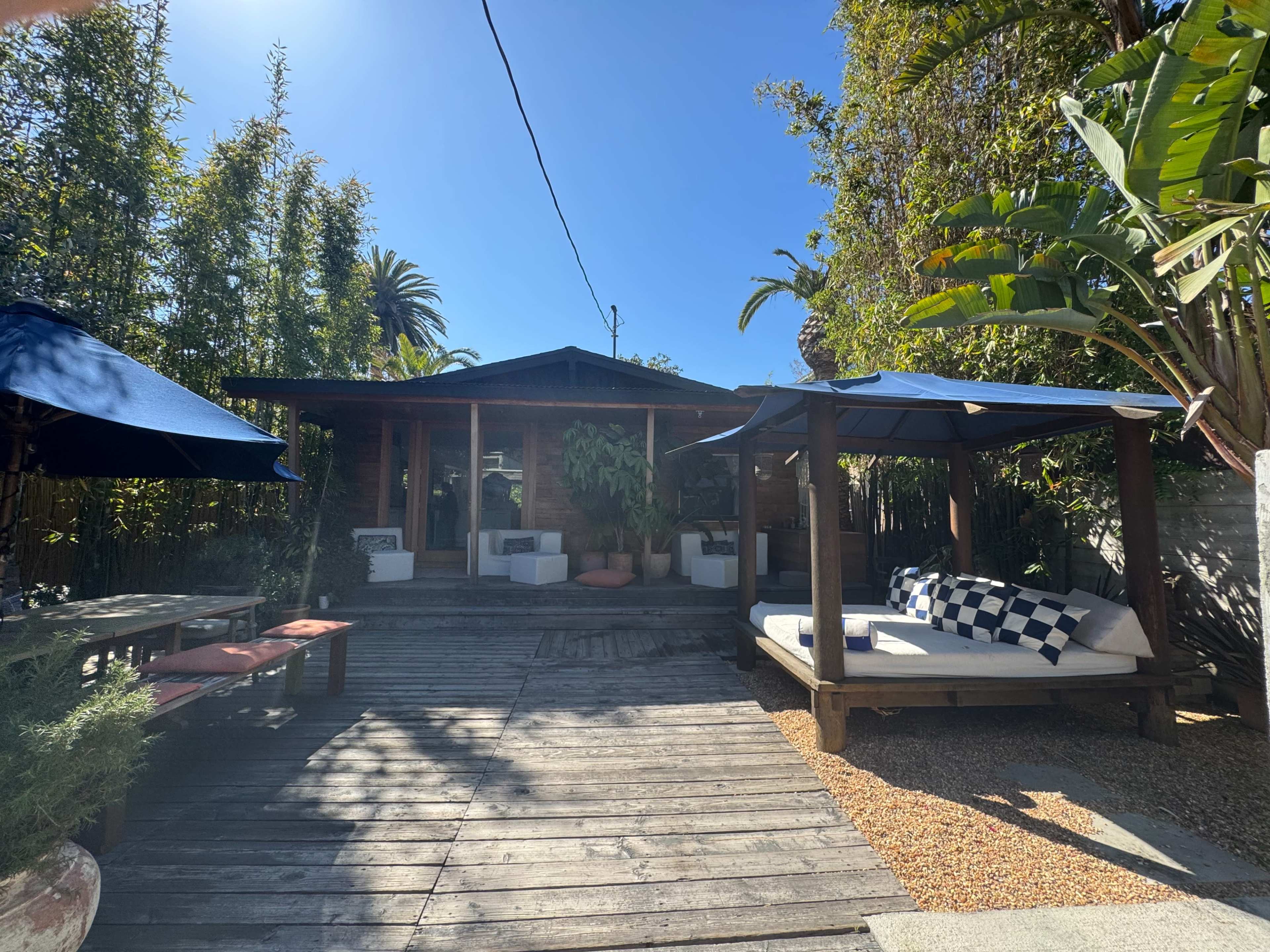 The image shows a wooden deck in a tranquil garden setting with a small wooden house and outdoor seating areas covered by blue umbrellas.