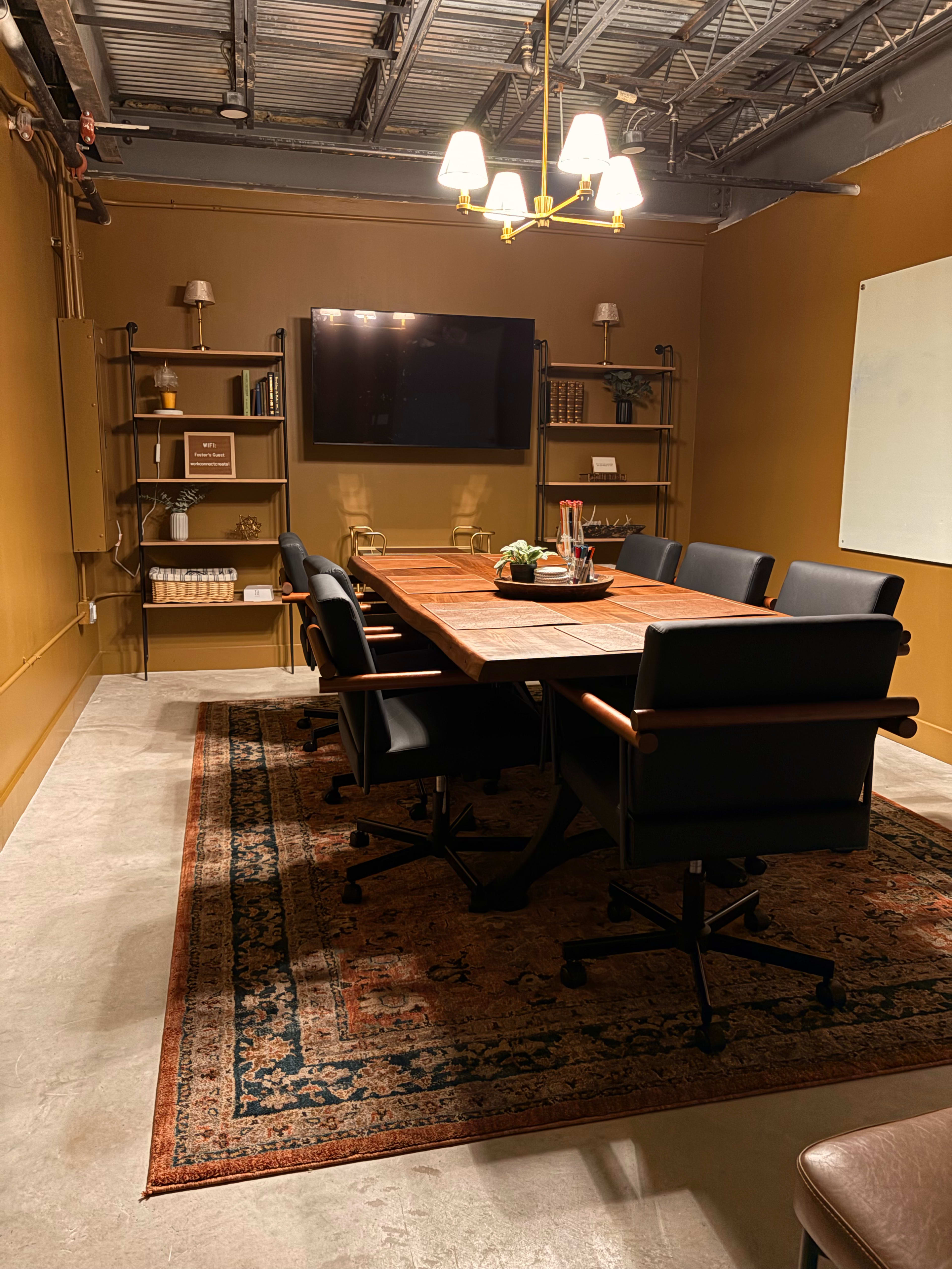 A conference room features a large wooden table surrounded by black chairs, with a television mounted on the wall and shelves filled with books and decor.