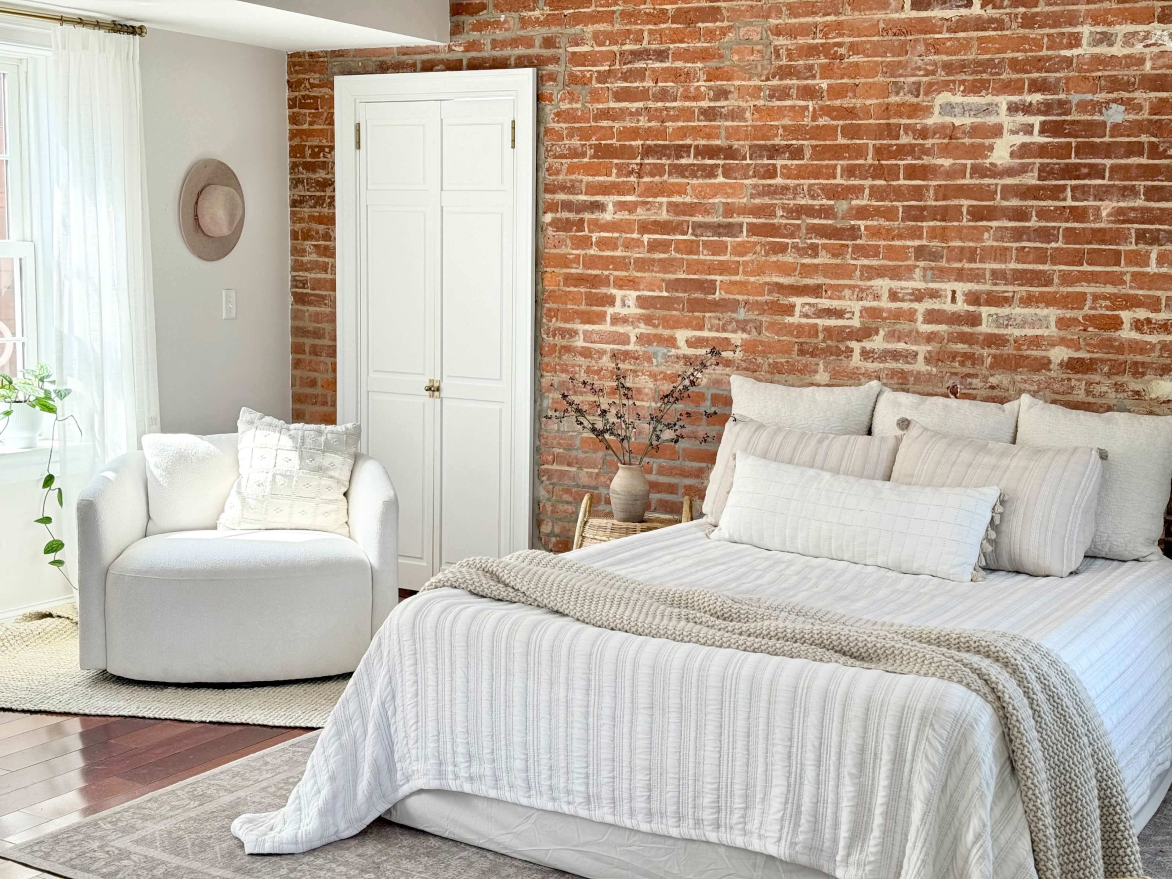 A cozy bedroom features a white bed with layered bedding and a round armchair, set against a brick accent wall.