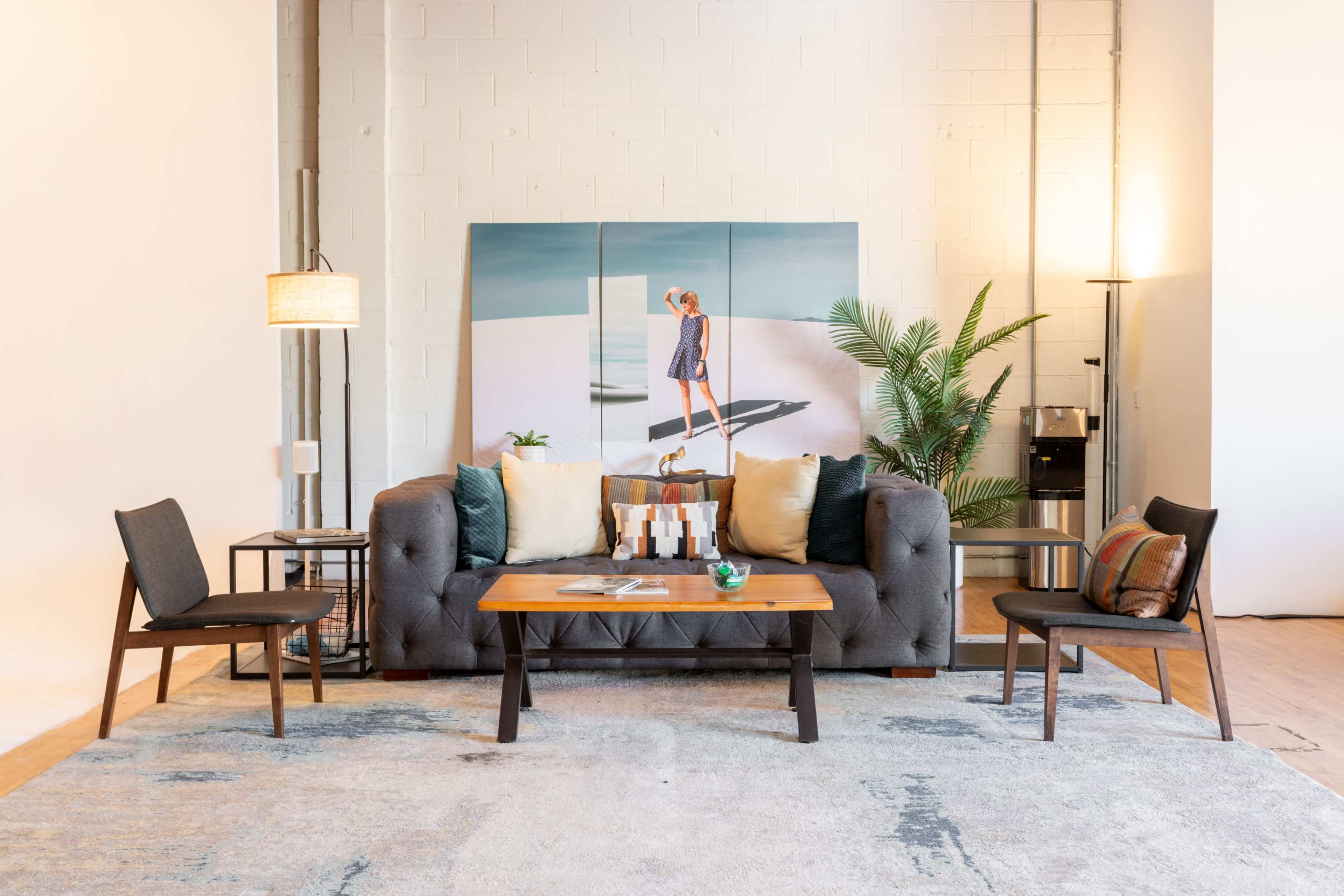 The image shows a styled living room featuring a dark gray couch, two chairs, a wooden coffee table, and a large artwork on the wall, accompanied by a floor lamp and a plant.