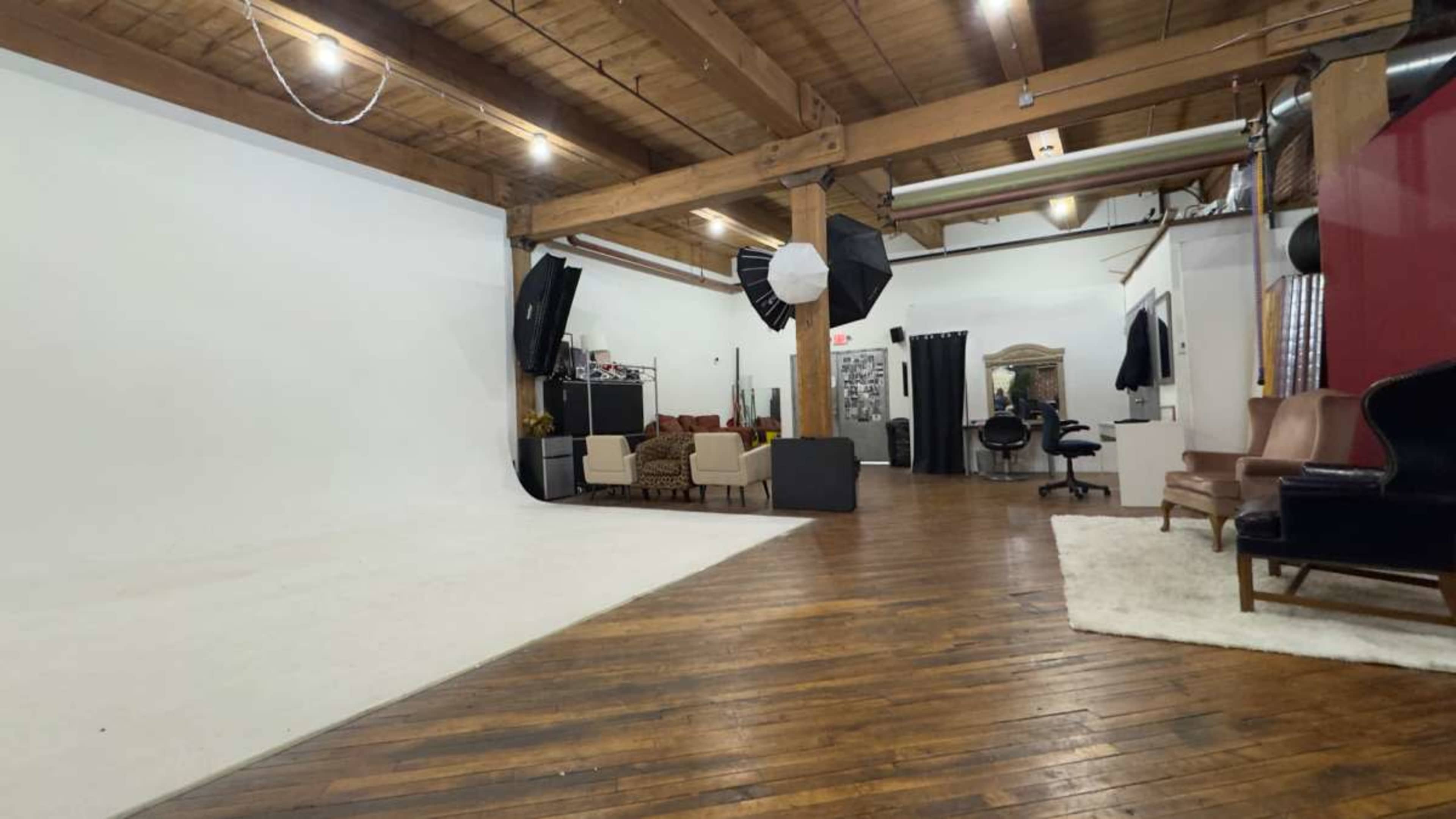 The image shows a spacious photography studio with a white backdrop, wooden floors, and various furniture pieces, including chairs and a small table.