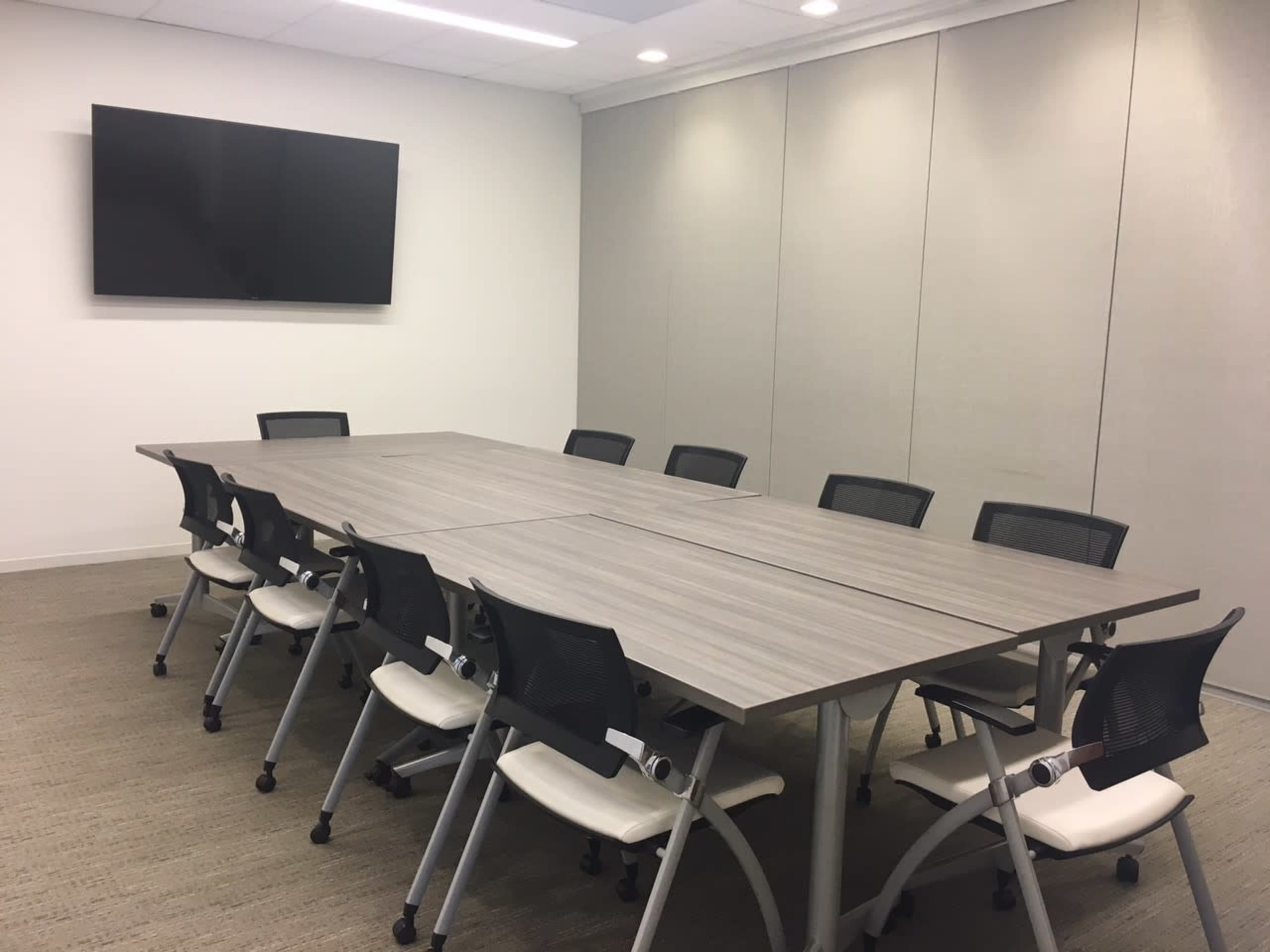 A spacious conference room features a large, rectangular table surrounded by eight chairs, with a wall-mounted television in the background.