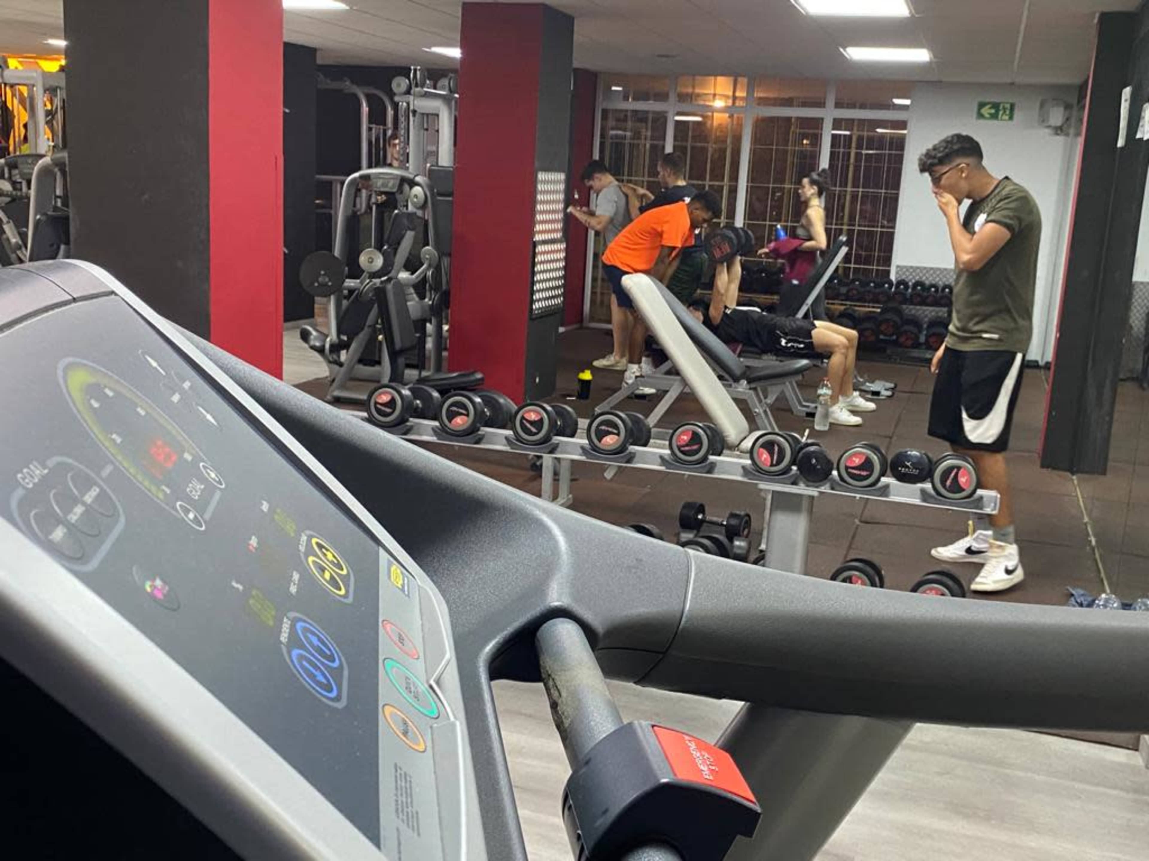 A view of a gym interior with various exercise equipment, including a treadmill in the foreground and several people engaged in workouts in the background.