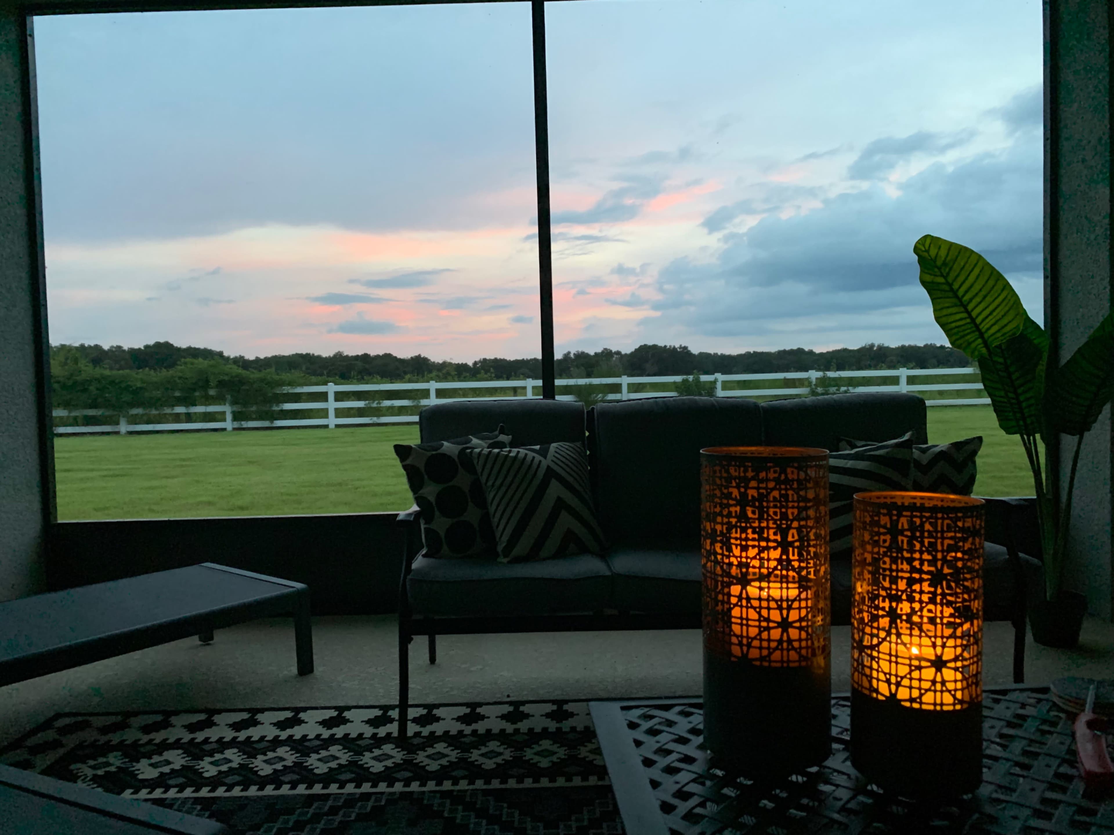 A cozy indoor seating area with two lit candles on a table, overlooking a grassy field and a colorful sky at dusk.