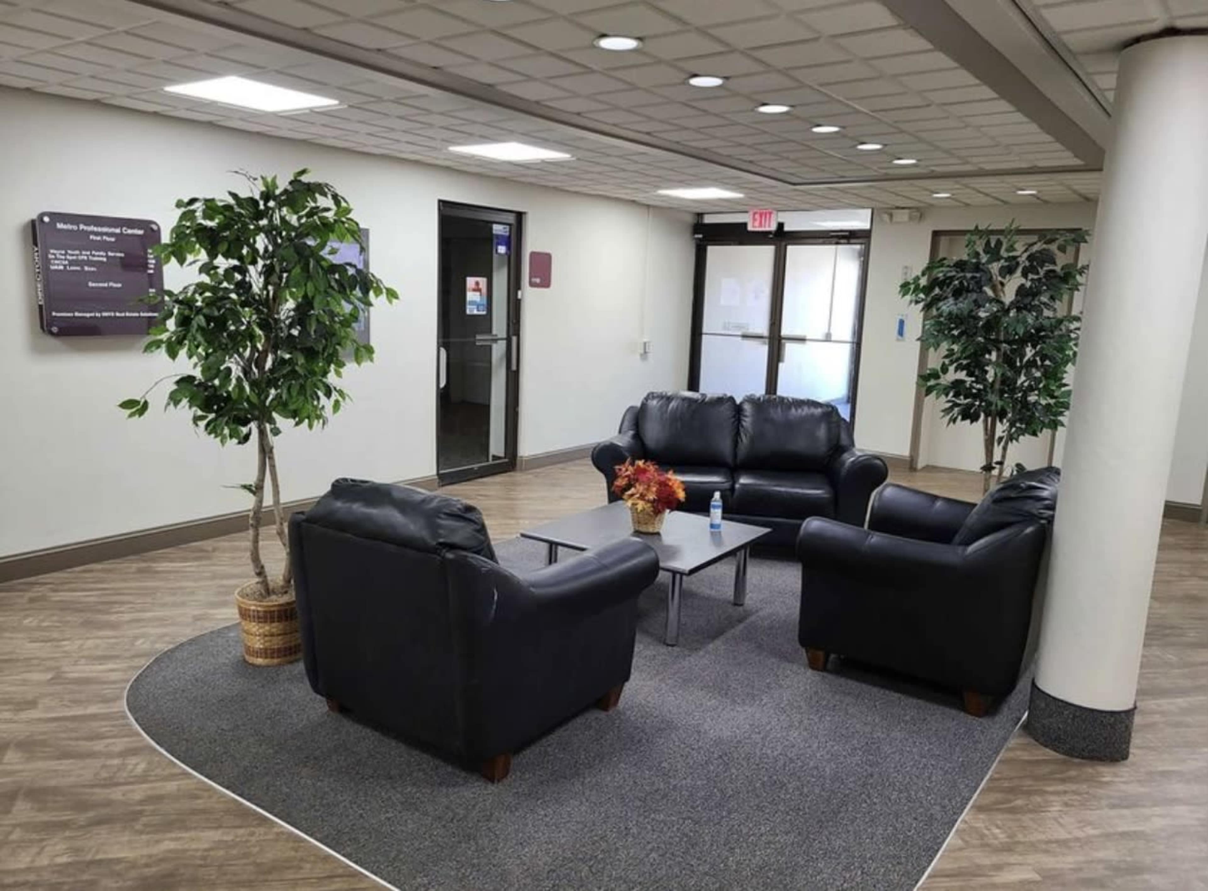 A waiting area with two black sofas, a glass coffee table, and two indoor trees, located in a well-lit hallway with doors leading to other rooms.