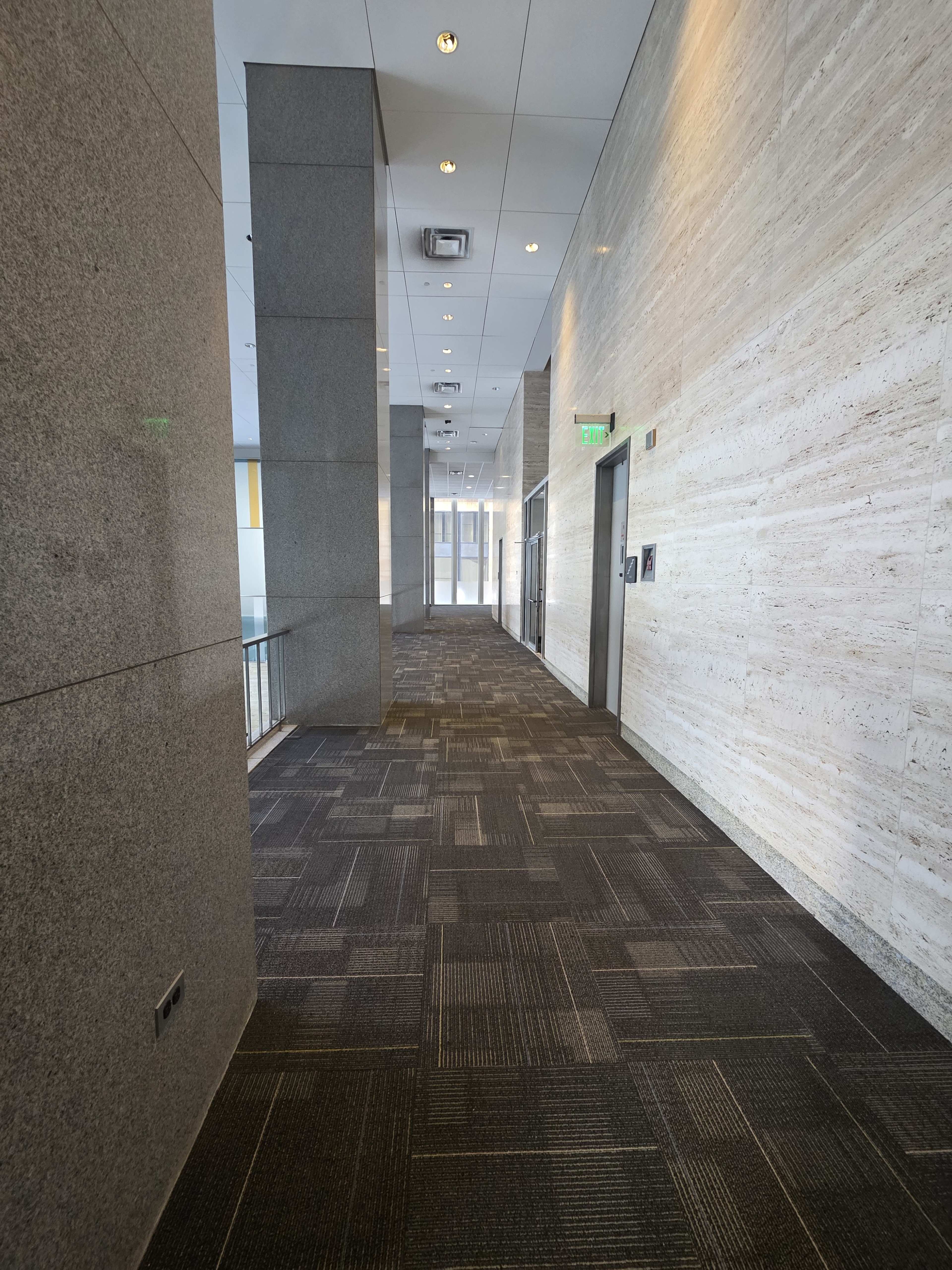 The image shows a long corridor with gray stone walls, carpeted flooring, and a door at the end, leading to a windowed area.