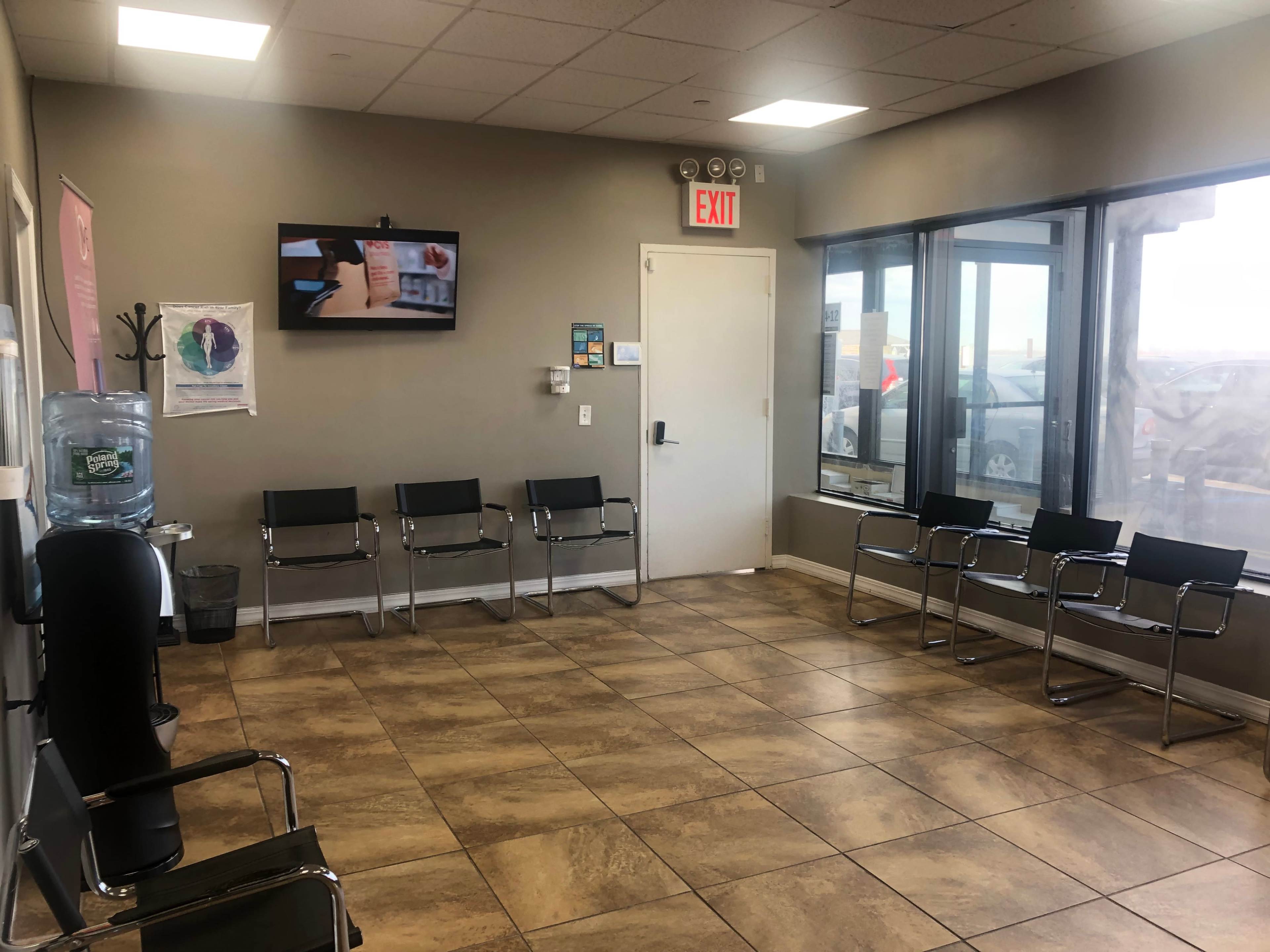 The image shows a waiting room with a row of metal chairs, a water cooler, a television on the wall, and large windows providing a view outside.