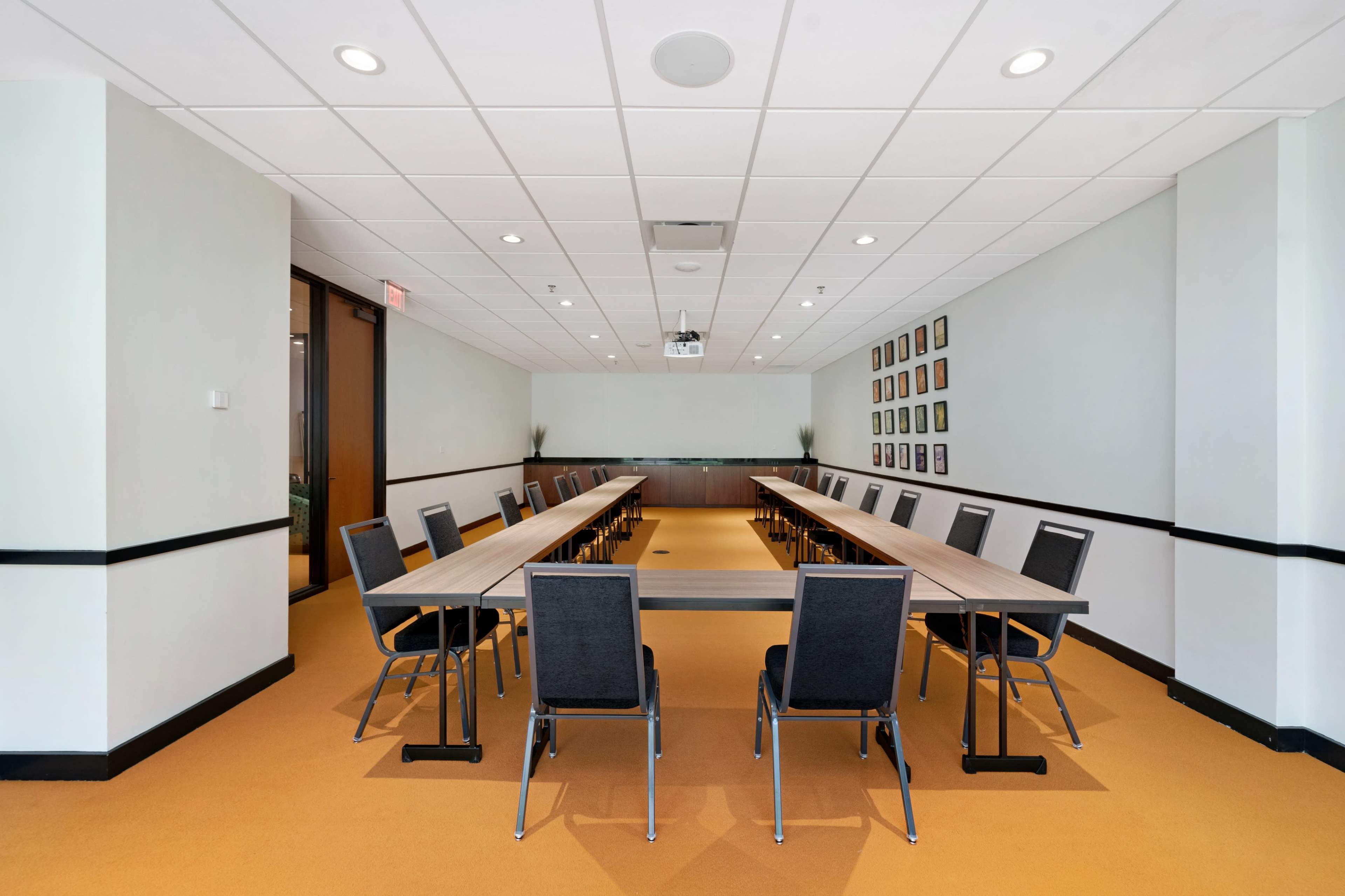 A spacious meeting room features a long table surrounded by chairs, with artwork displayed on the walls and a projector mounted on the ceiling.