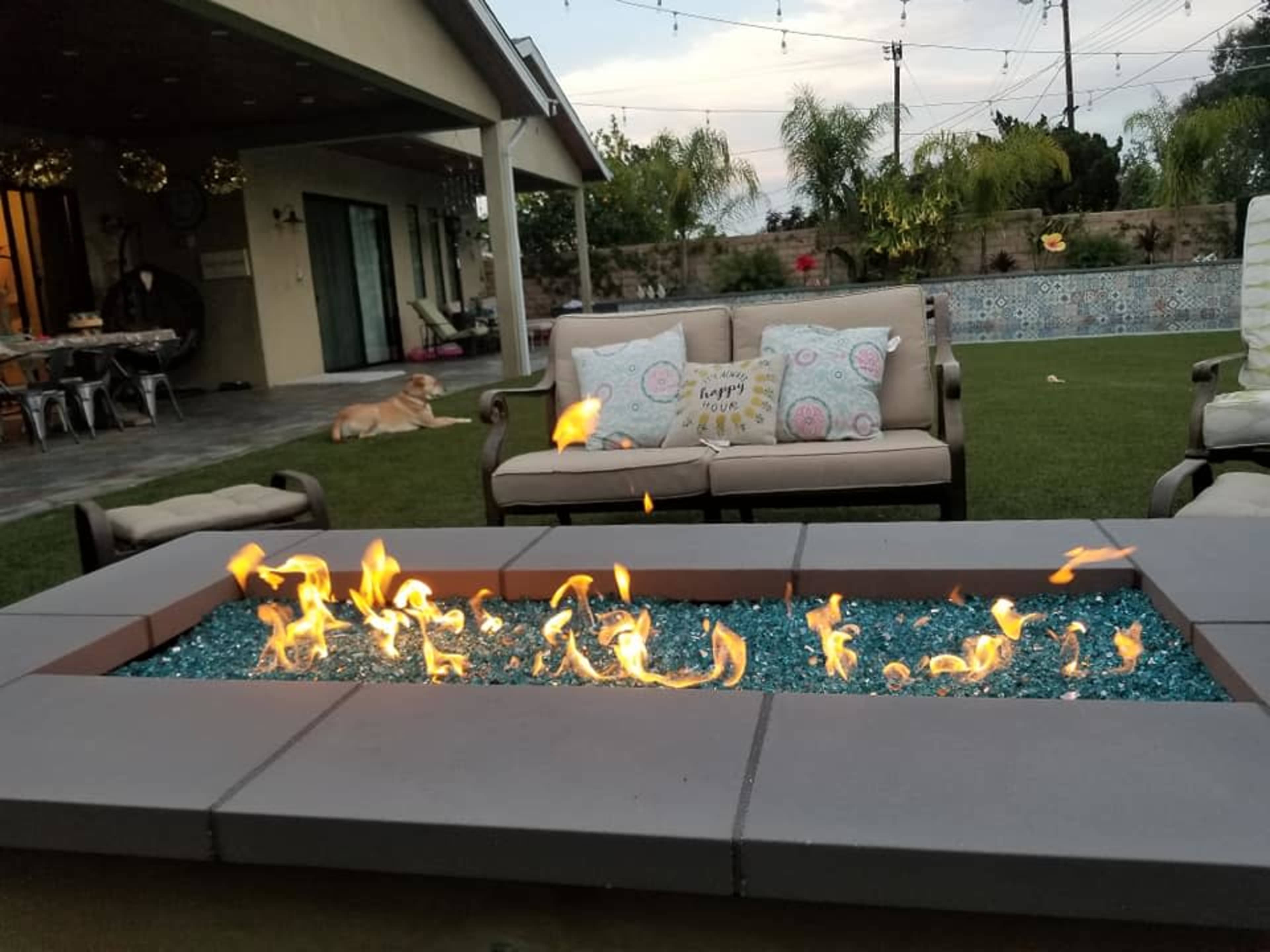 A fire pit with blue glass stones burns in the foreground, while two sofas and a dog are visible in a backyard setting.