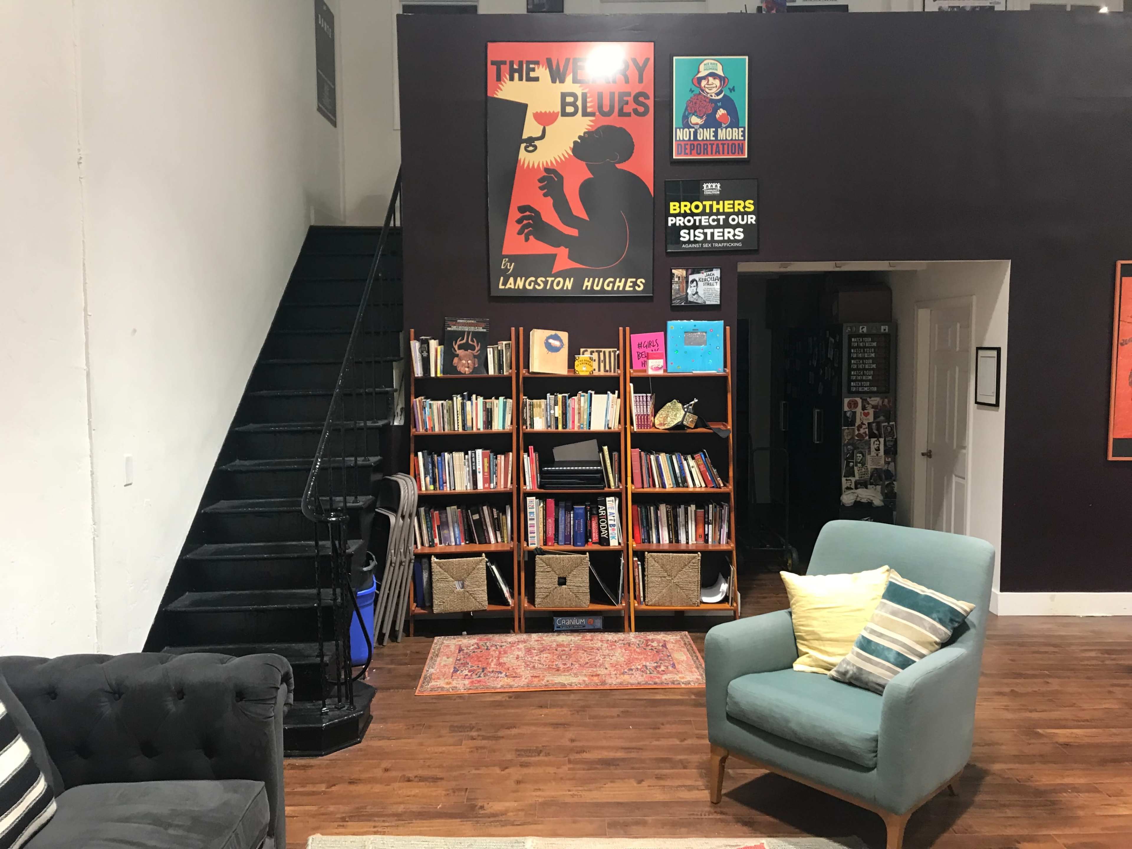 The image shows a room with a staircase on the left, a bookshelf filled with books in the center, and a seating area featuring a blue armchair and a dark couch.