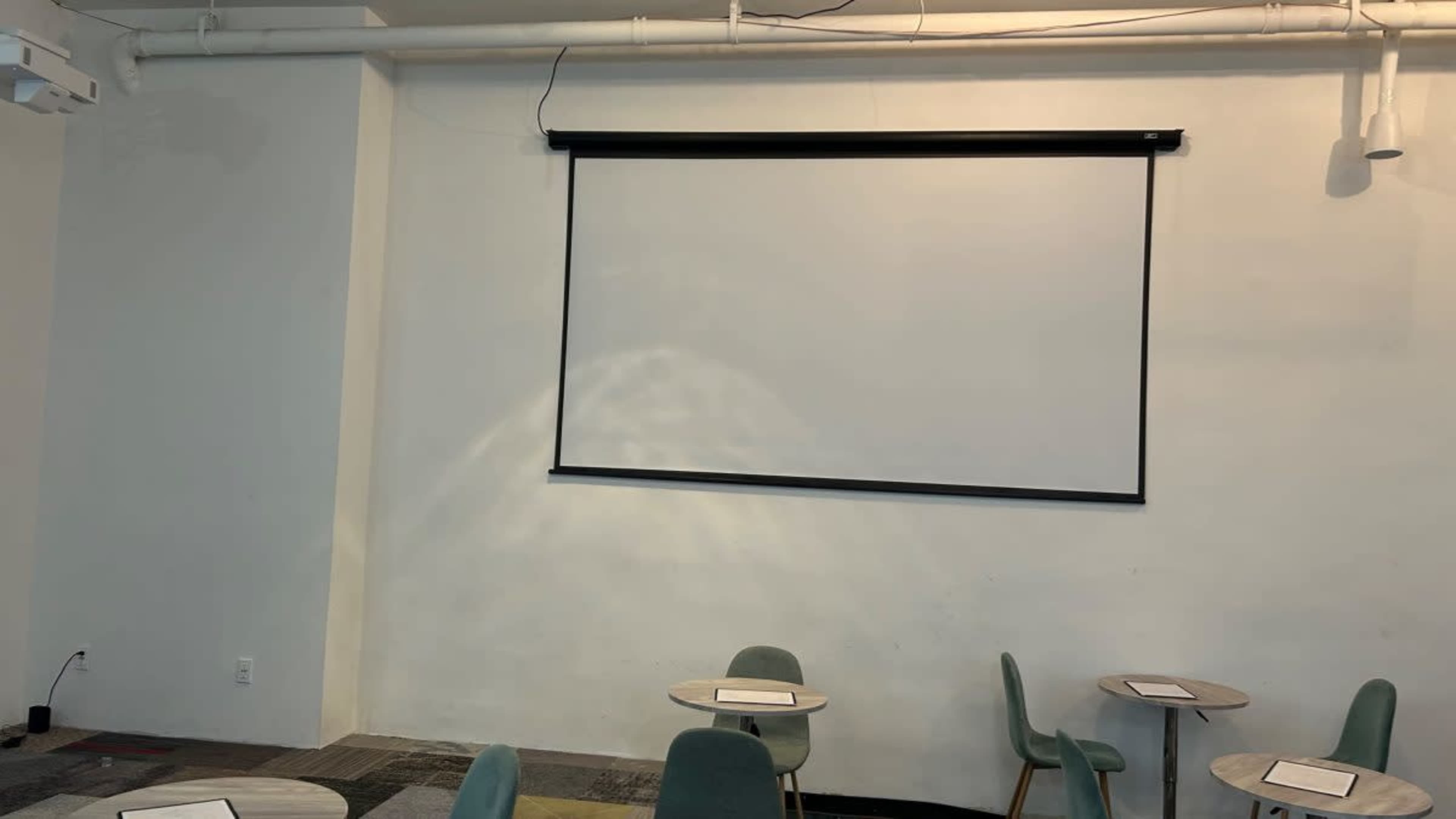 A blank projection screen on a white wall with two round tables and green chairs in a room.