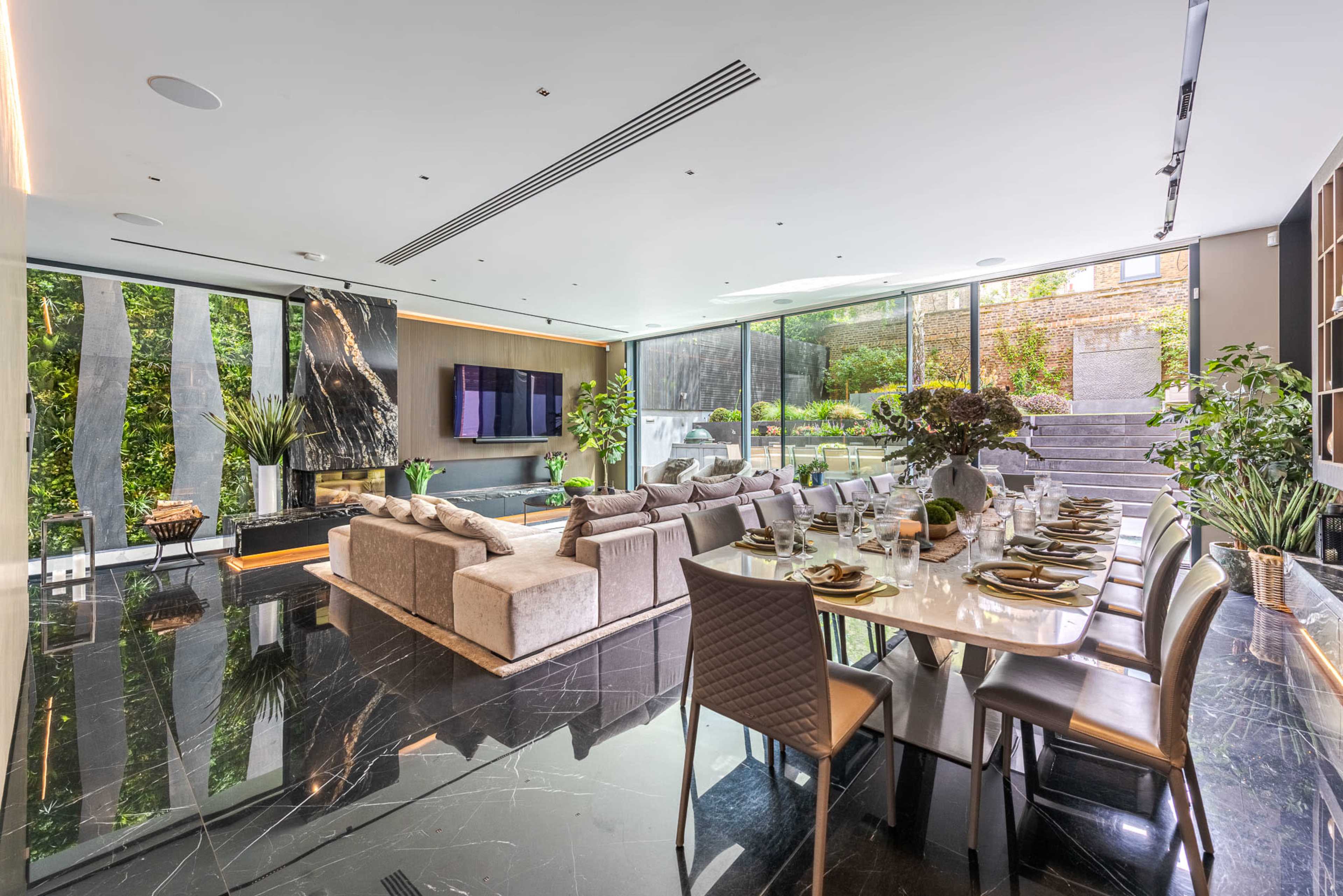 A modern living and dining area features sleek furniture, dark marble flooring, and large windows overlooking a garden.