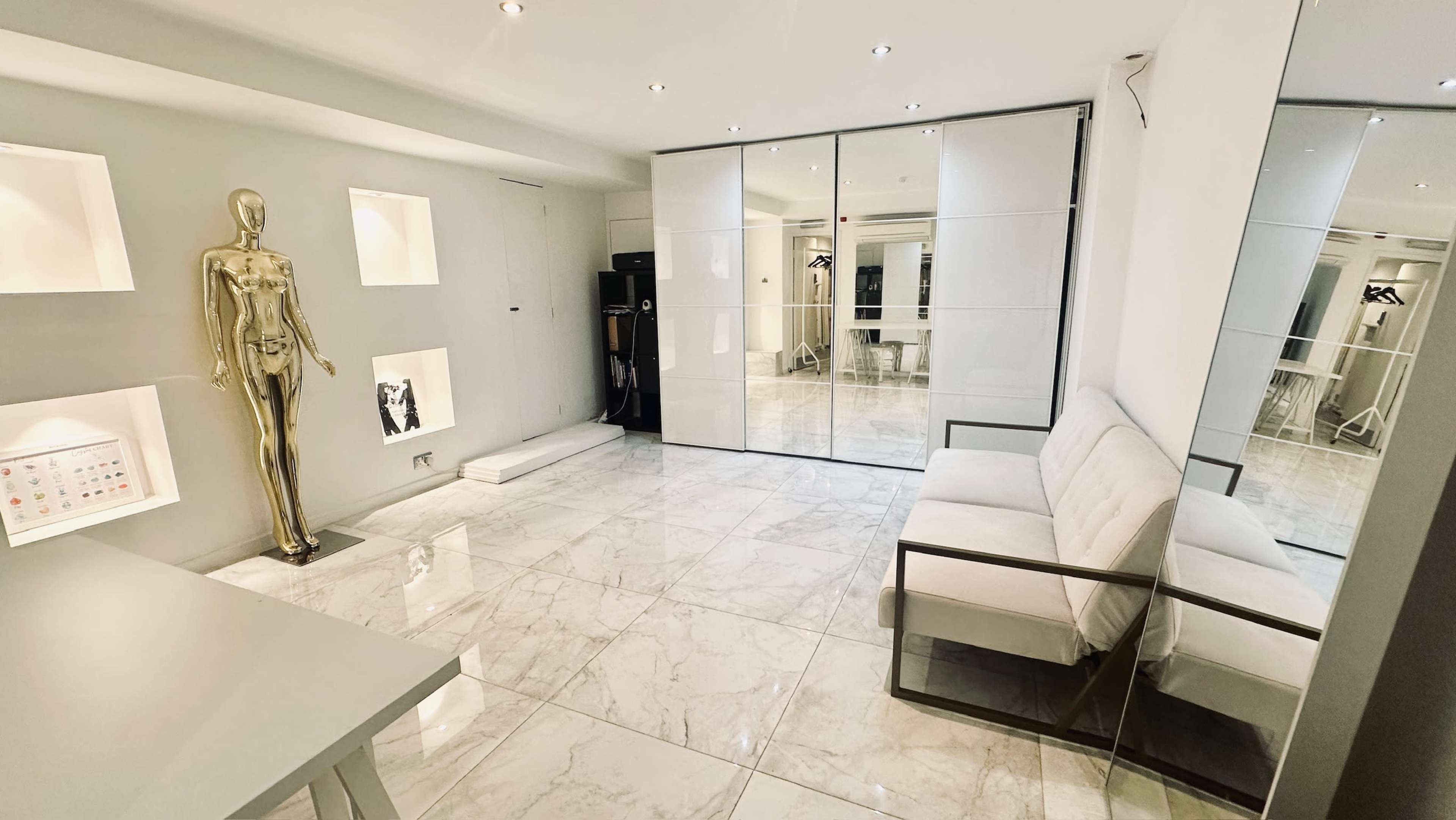 The image shows a modern, minimalist room with marble flooring, large mirrored panels, and a gold mannequin next to a white couch.