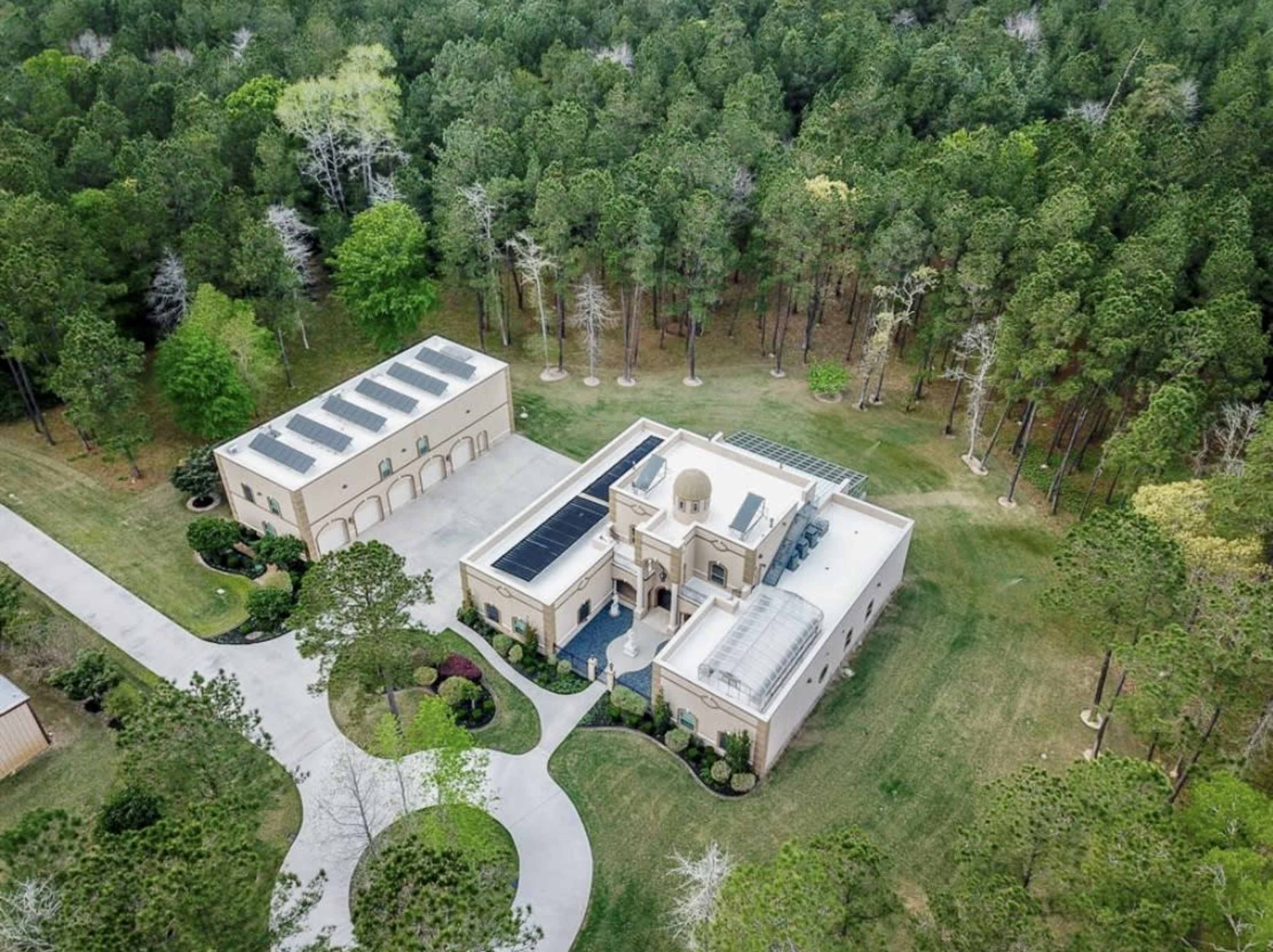 An aerial view shows a large, modern home surrounded by dense trees and a well-maintained driveway leading to a garage.