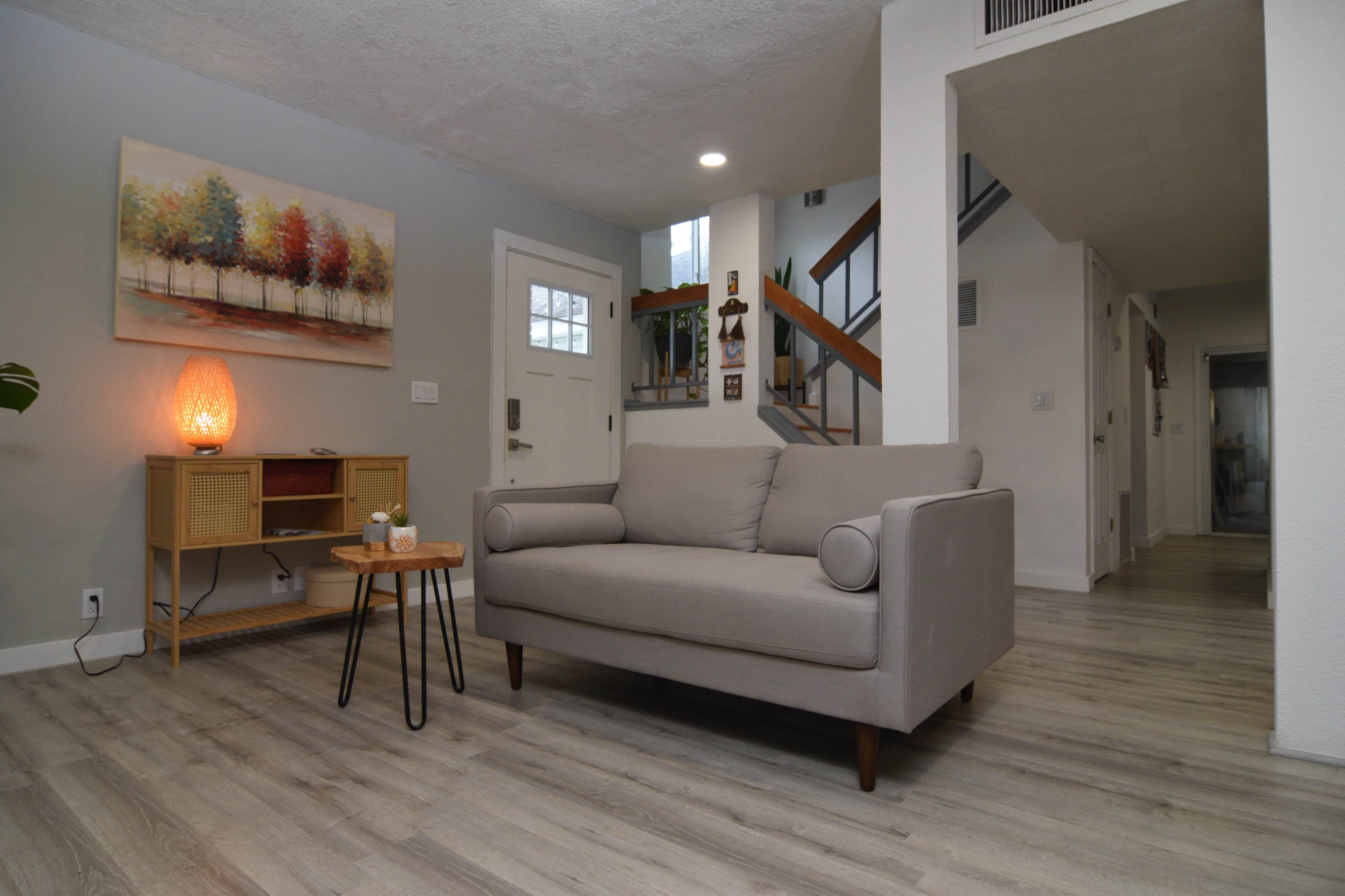 A living room featuring a light gray sofa, a small round wooden table, and a wall artwork of trees, with a staircase visible in the background.