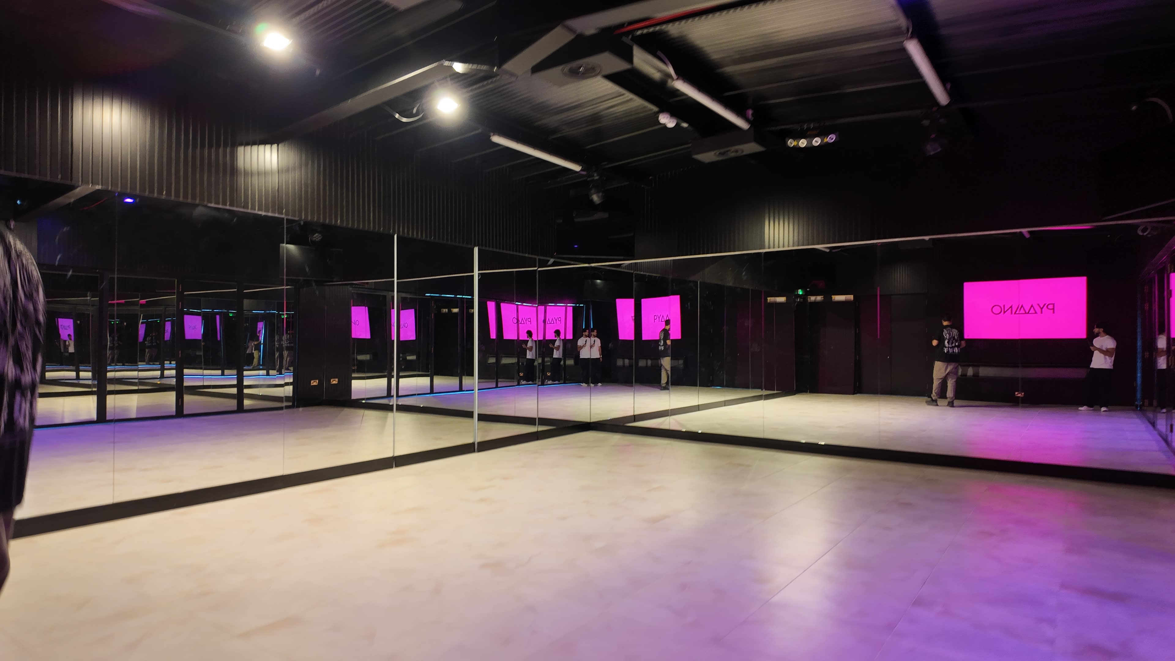 The image shows a mirrored dance studio with dark walls and pink lighting, featuring a spacious floor and multiple reflections of the interior.