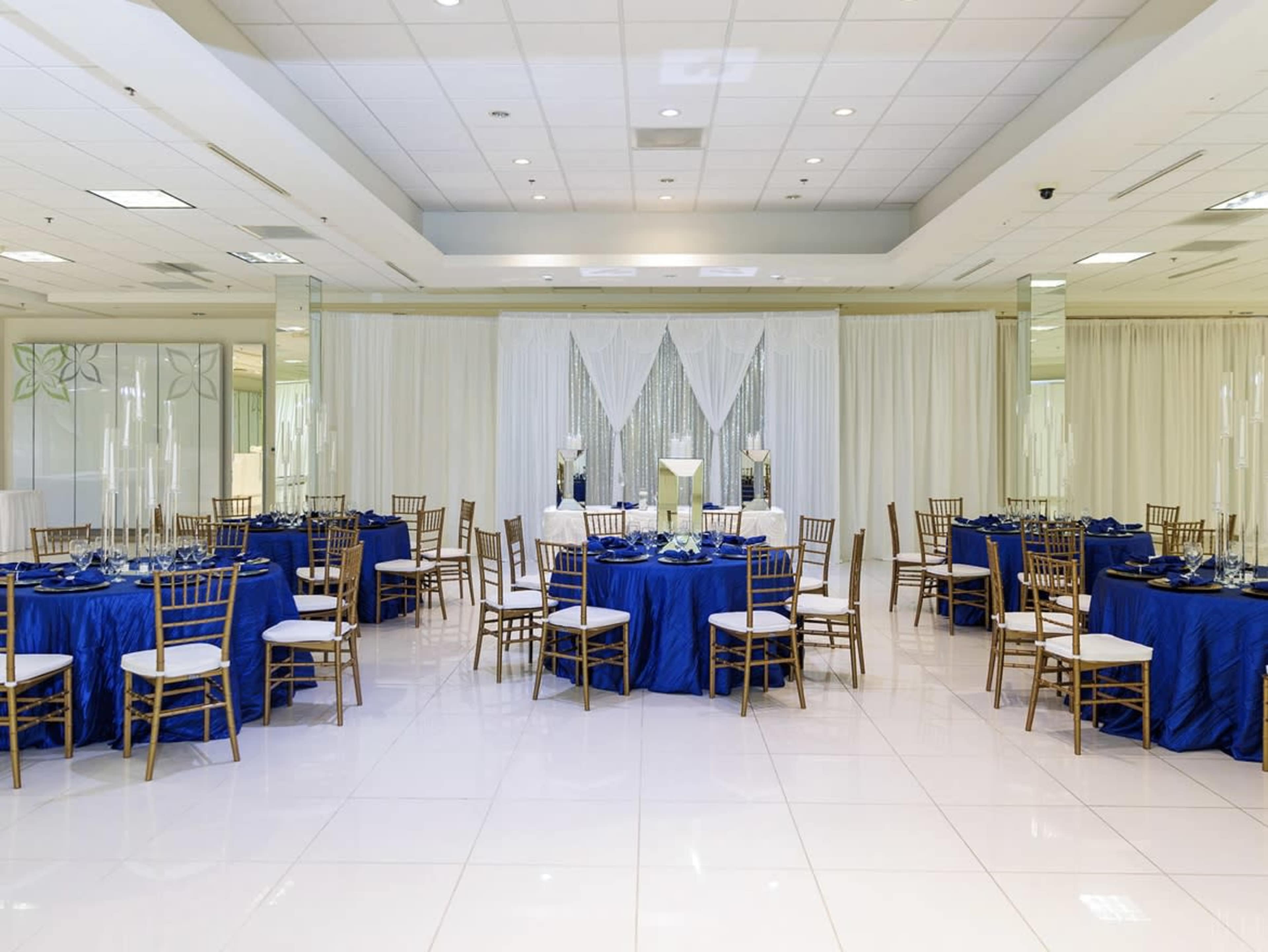 The image shows a banquet hall set up with round tables covered in blue tablecloths and gold chairs arranged around them, with white curtains and decorative elements in the background.