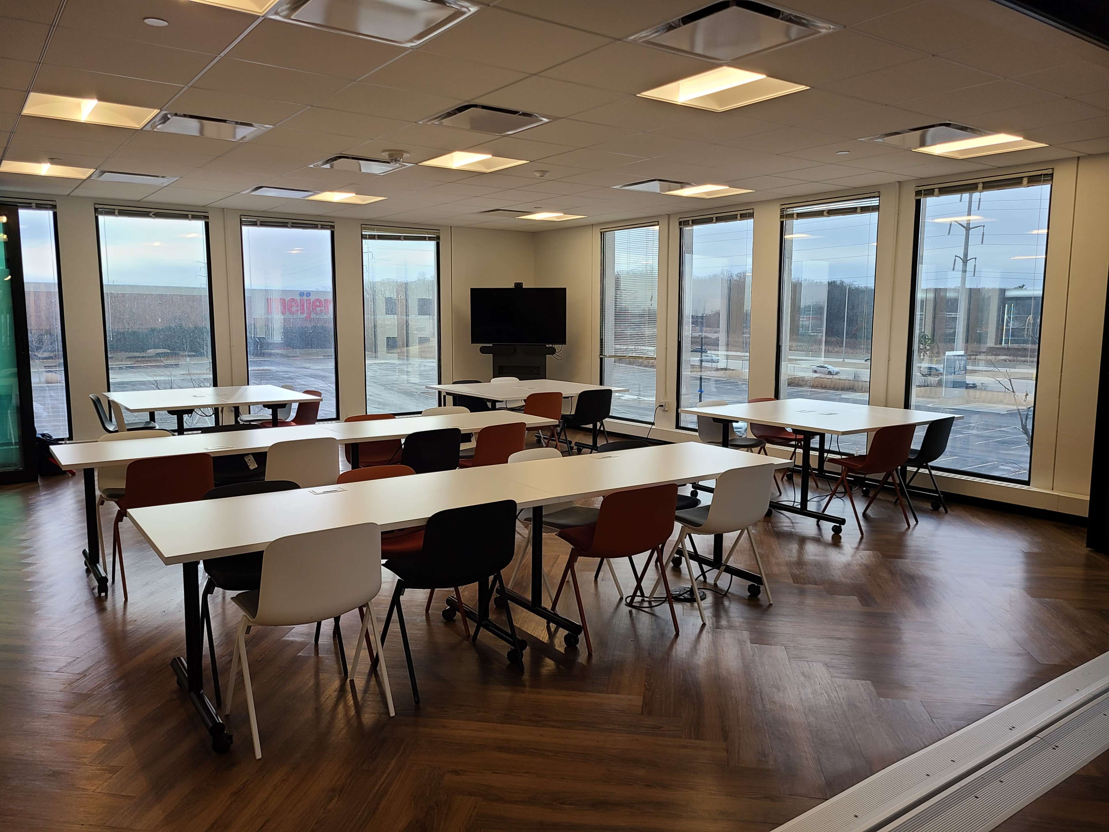 The image shows a modern conference room with several tables and chairs arranged around the space, featuring large windows that provide a view of the outside.