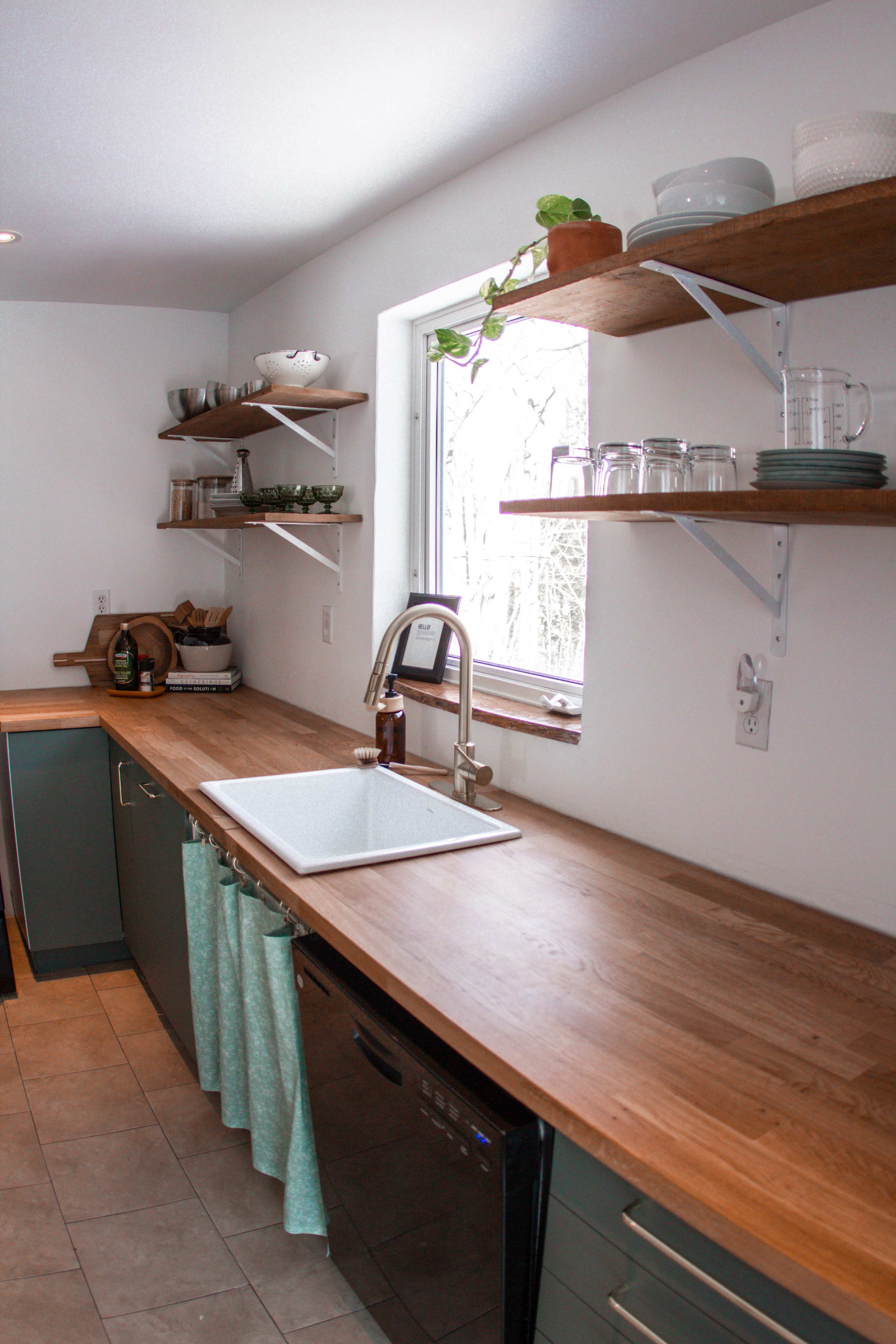 A modern kitchen features a long wooden countertop with a sink, shelves above holding dishes and plants, and a window letting in natural light.
