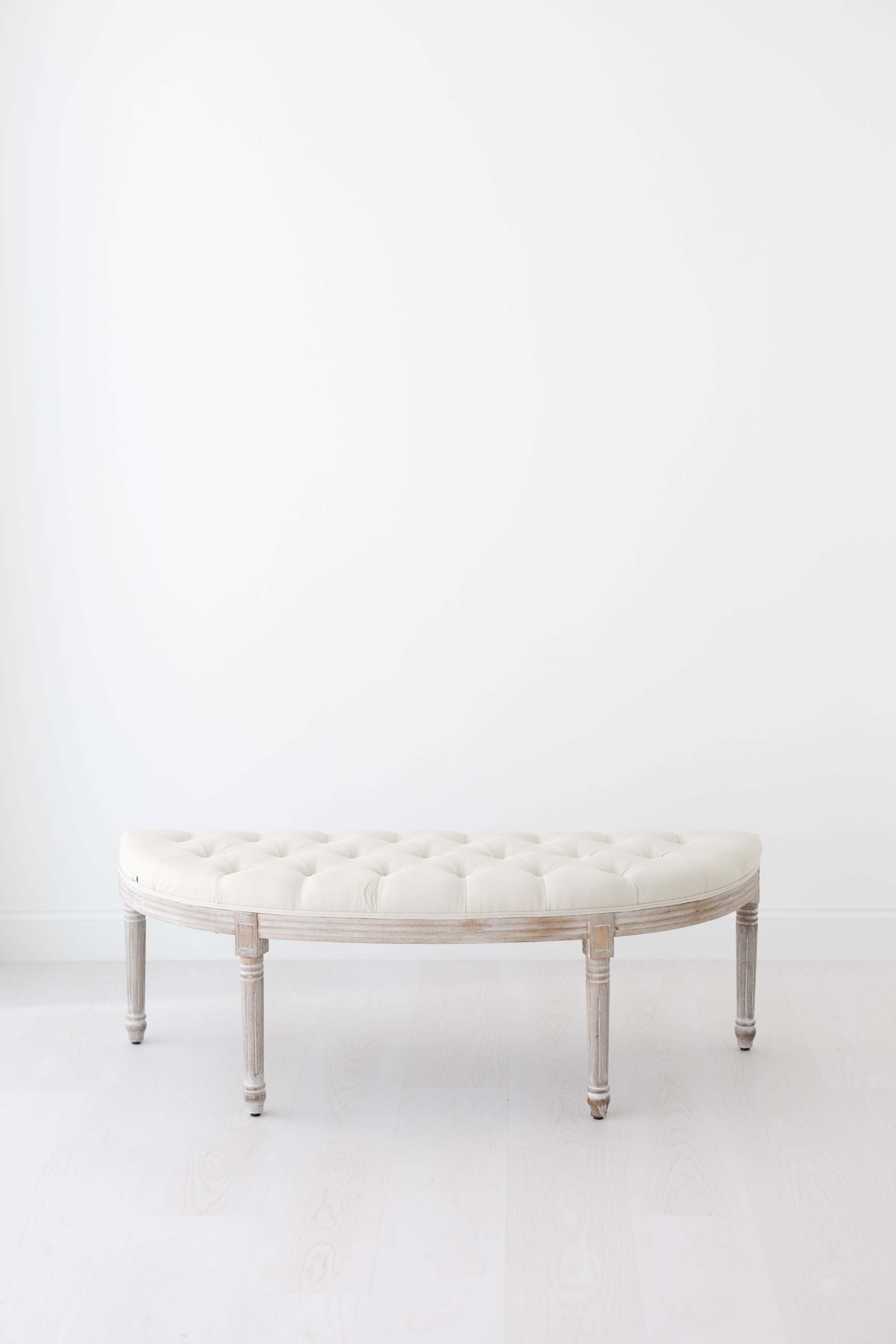 A tufted bench with a curved shape and light-colored fabric is placed against a plain white wall.