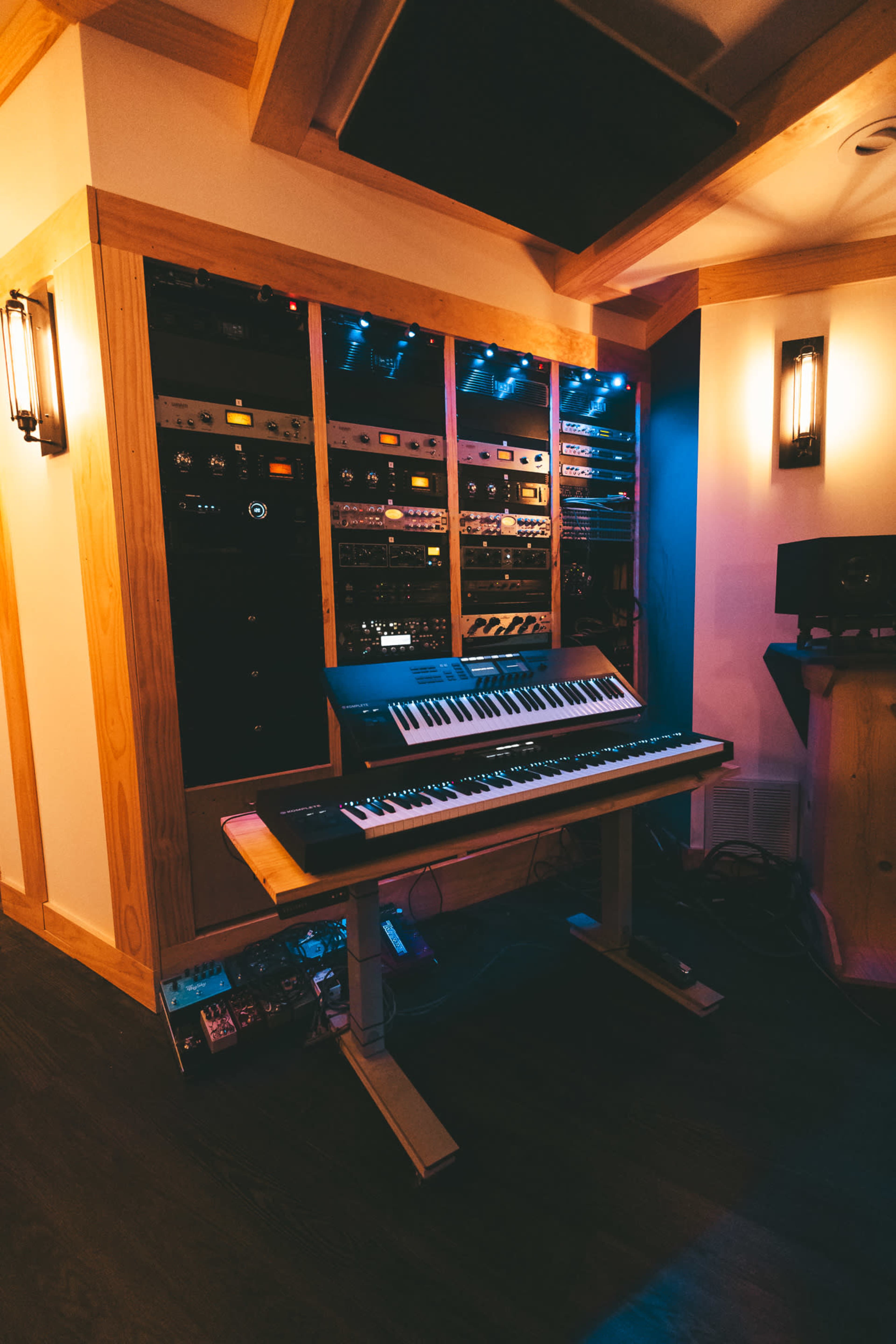 The image shows a music production studio with a keyboard on a stand in front of a wall of audio equipment and racks illuminated by blue lights.