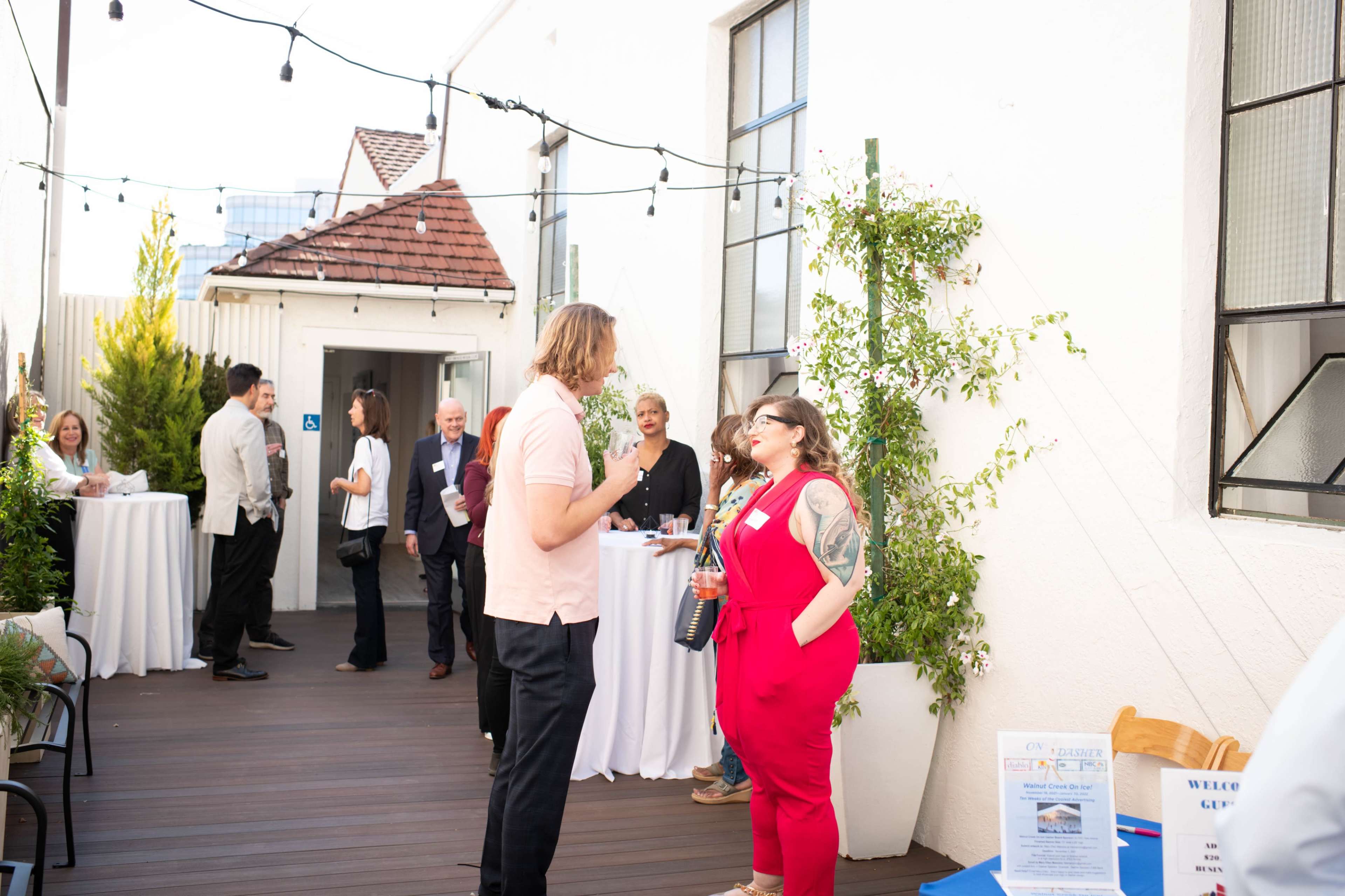A group of people engages in conversation at an outdoor event with string lights overhead and refreshments on display.