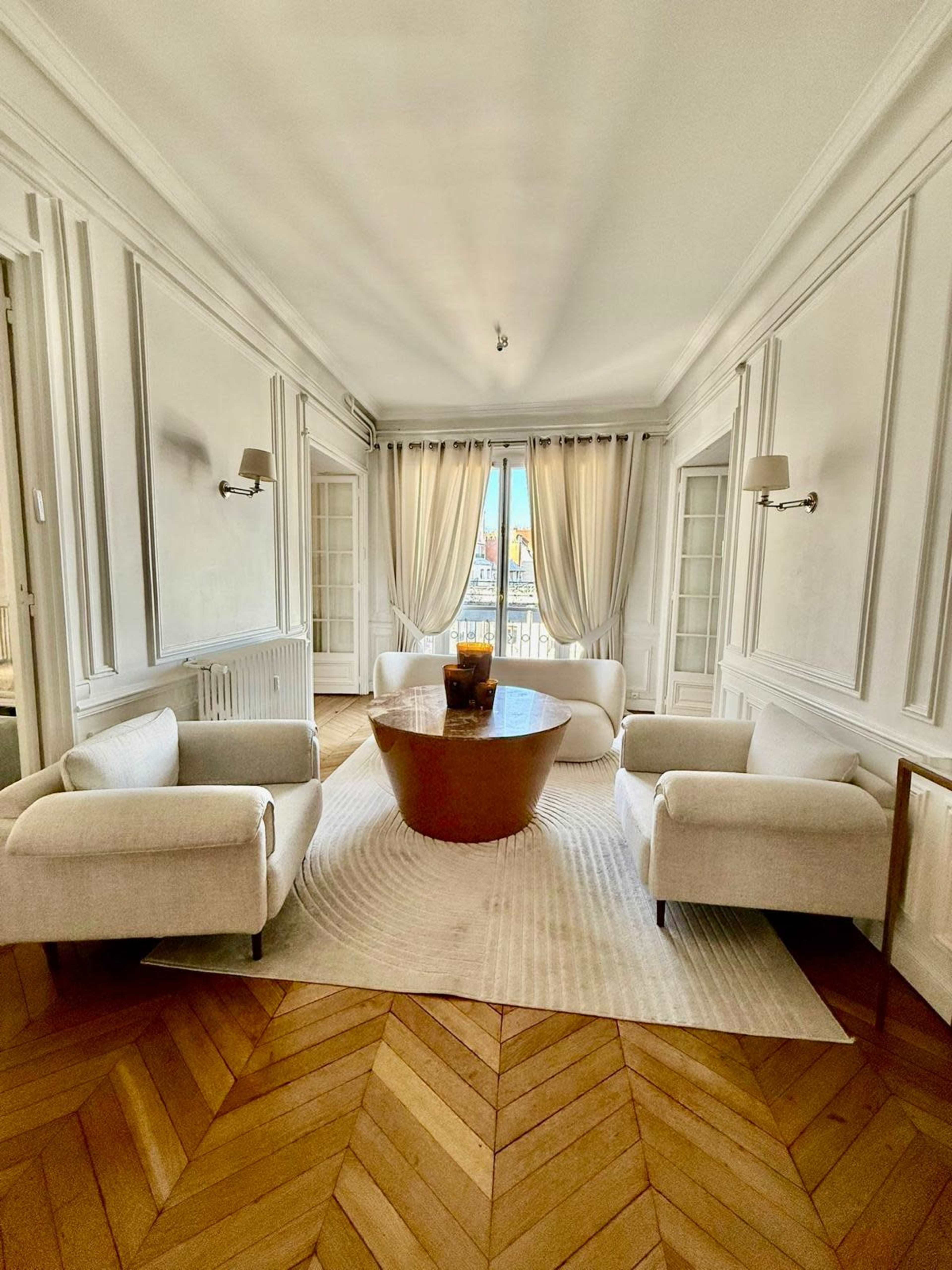 The room features a round table surrounded by two white armchairs on a light-colored rug, with large windows draped in curtains allowing natural light to enter.