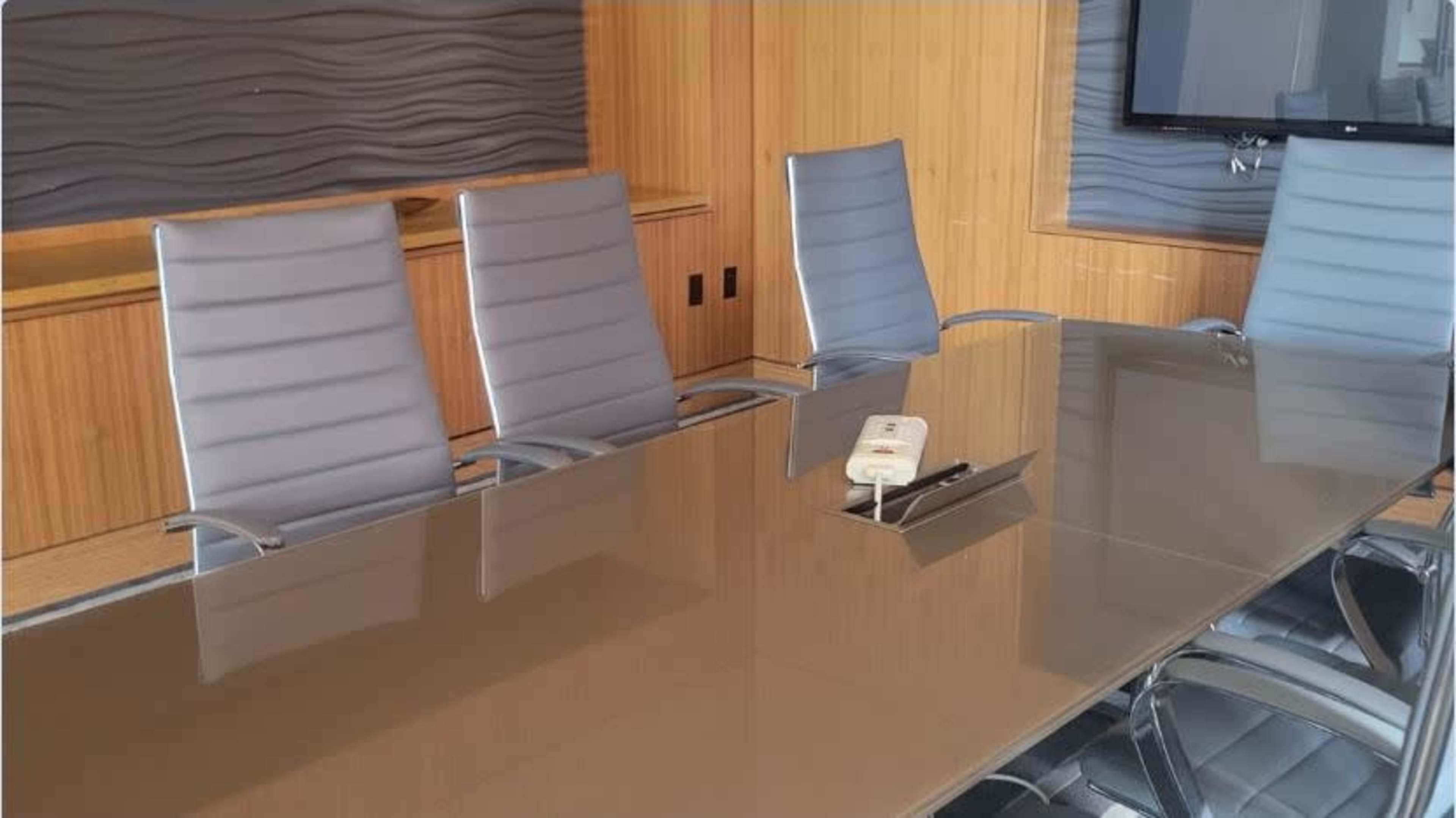 A conference room features a long glass table surrounded by multiple gray chairs and a wall-mounted television.