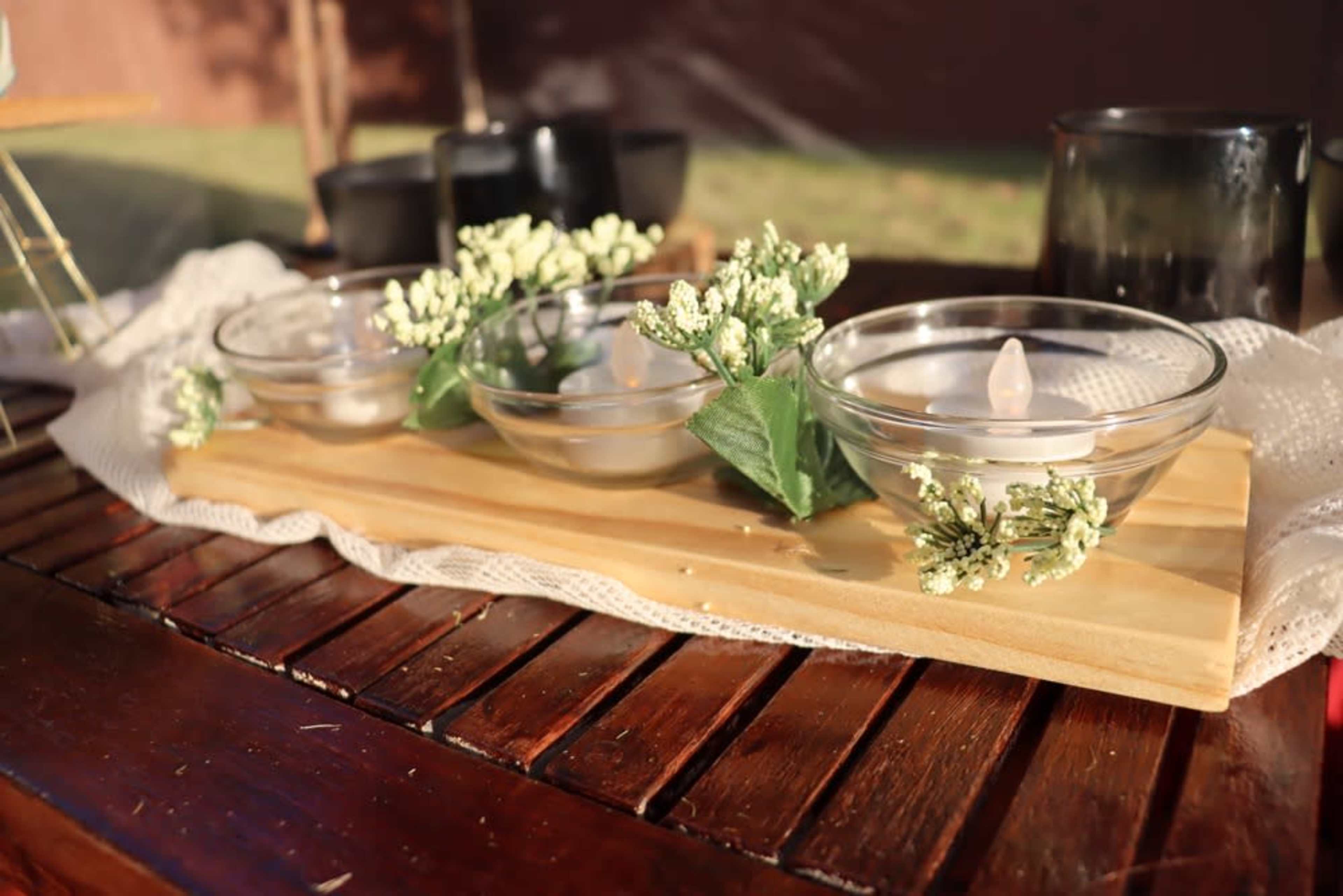 The image shows a wooden serving tray with three glass bowls containing candles, surrounded by faux greenery, placed on a wooden table.