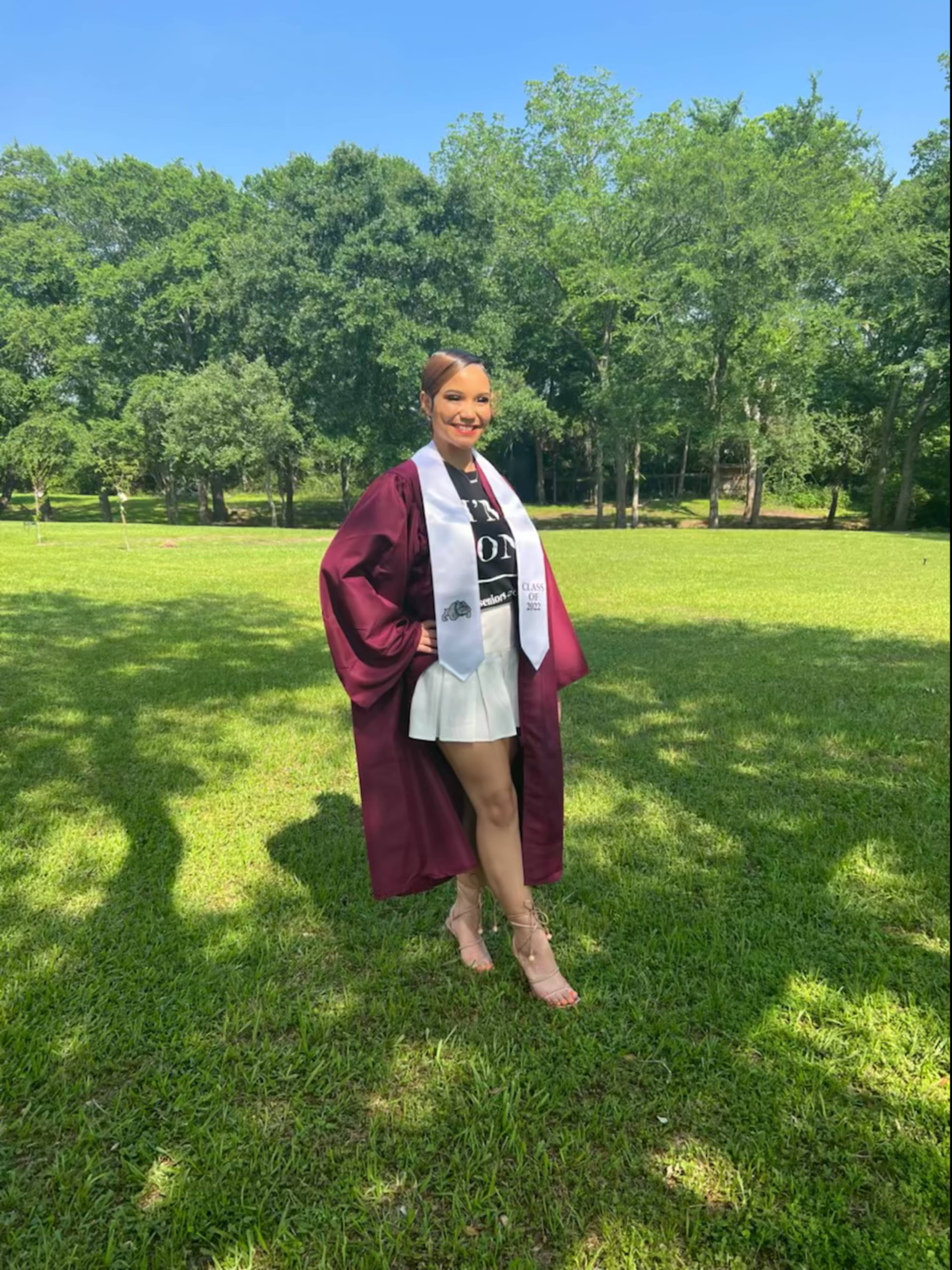 A graduate in a maroon robe and sash stands in a grassy field with trees in the background.