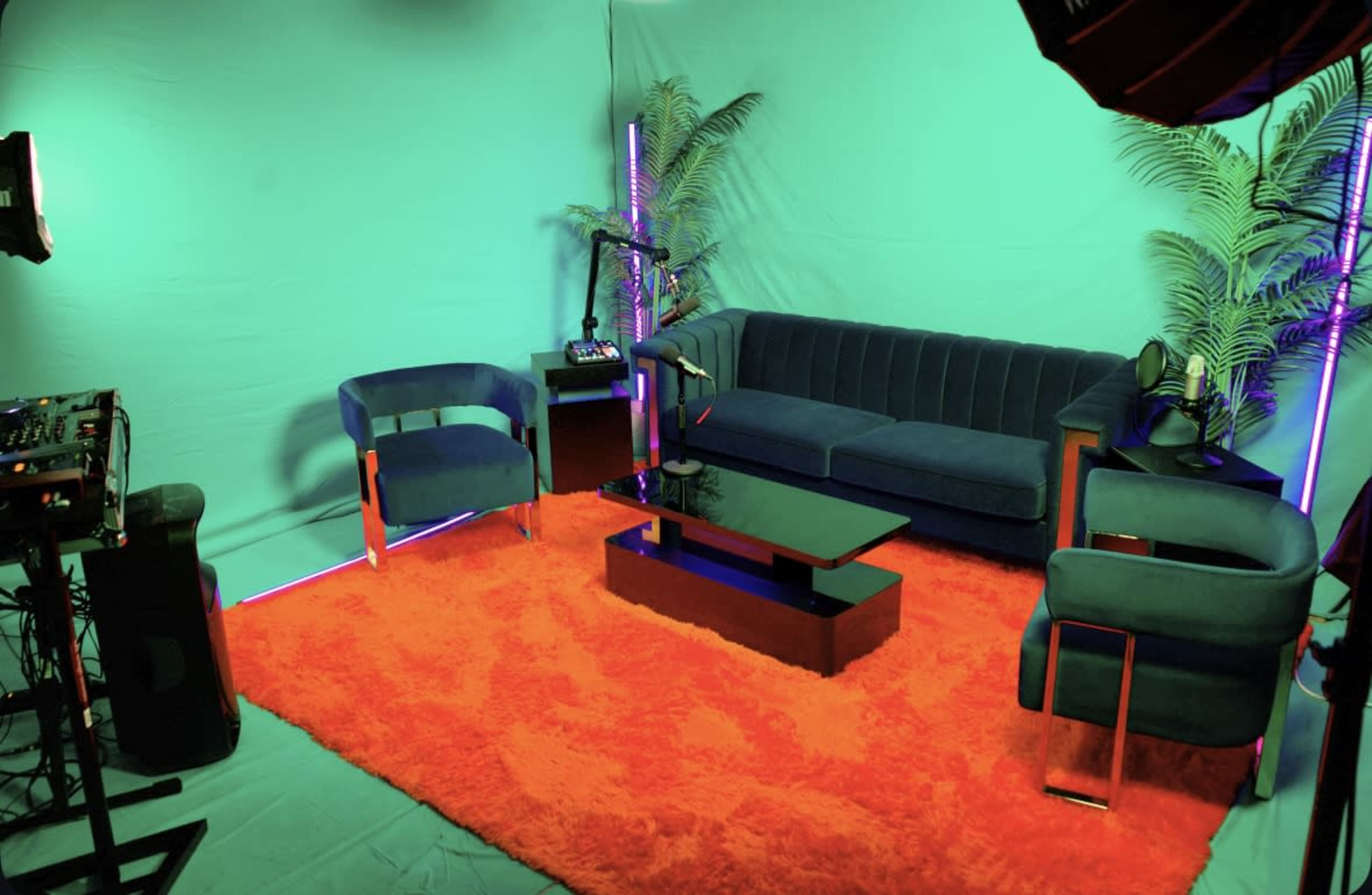 The image shows a modern sitting area featuring a blue couch, two blue chairs, and a black coffee table, set against a green backdrop with an orange rug and decorative plants.