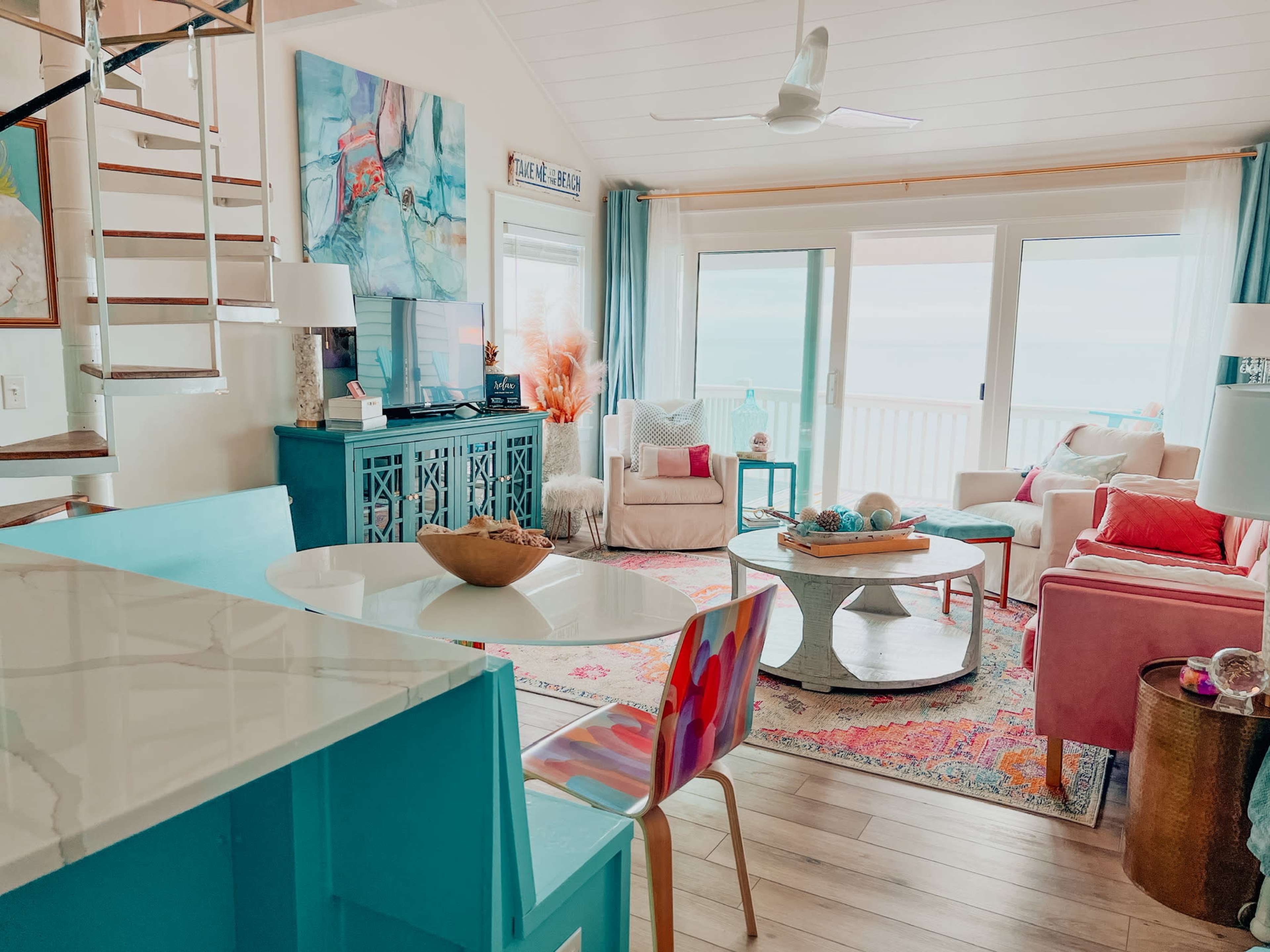 A bright living area with a turquoise cabinet, pink chairs, and a large window overlooking a beach view.
