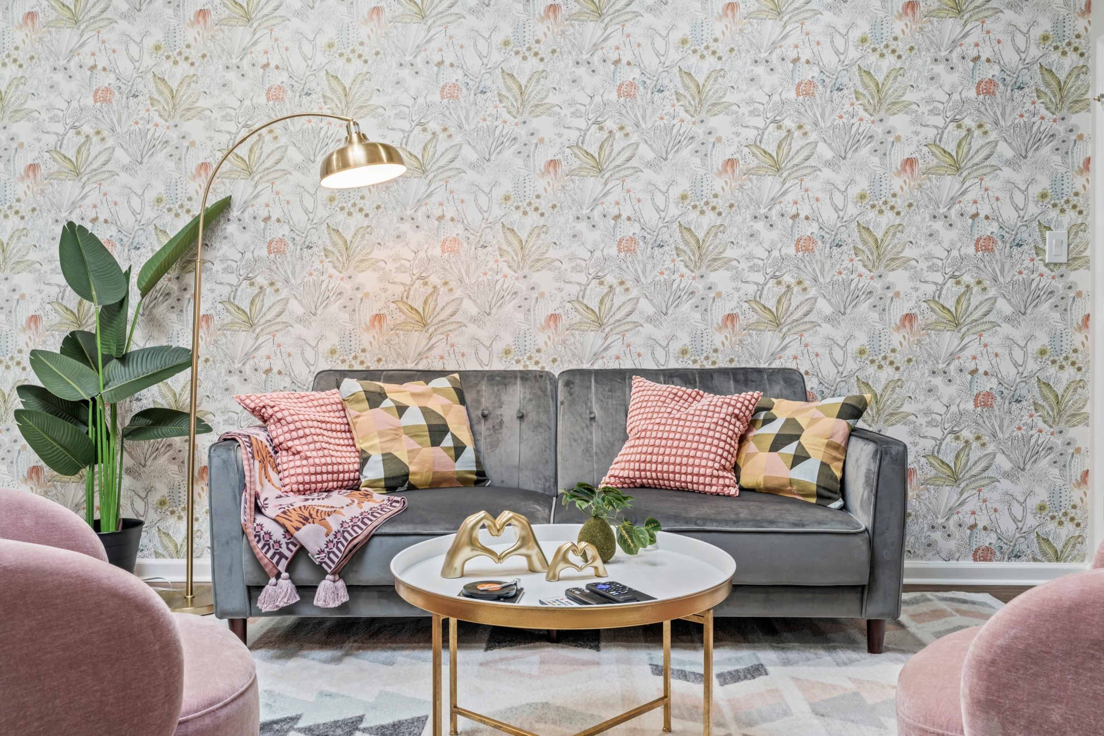 The image shows a living room with a gray sofa adorned with patterned cushions, a round coffee table, and decorative plants under a floral wallpaper.