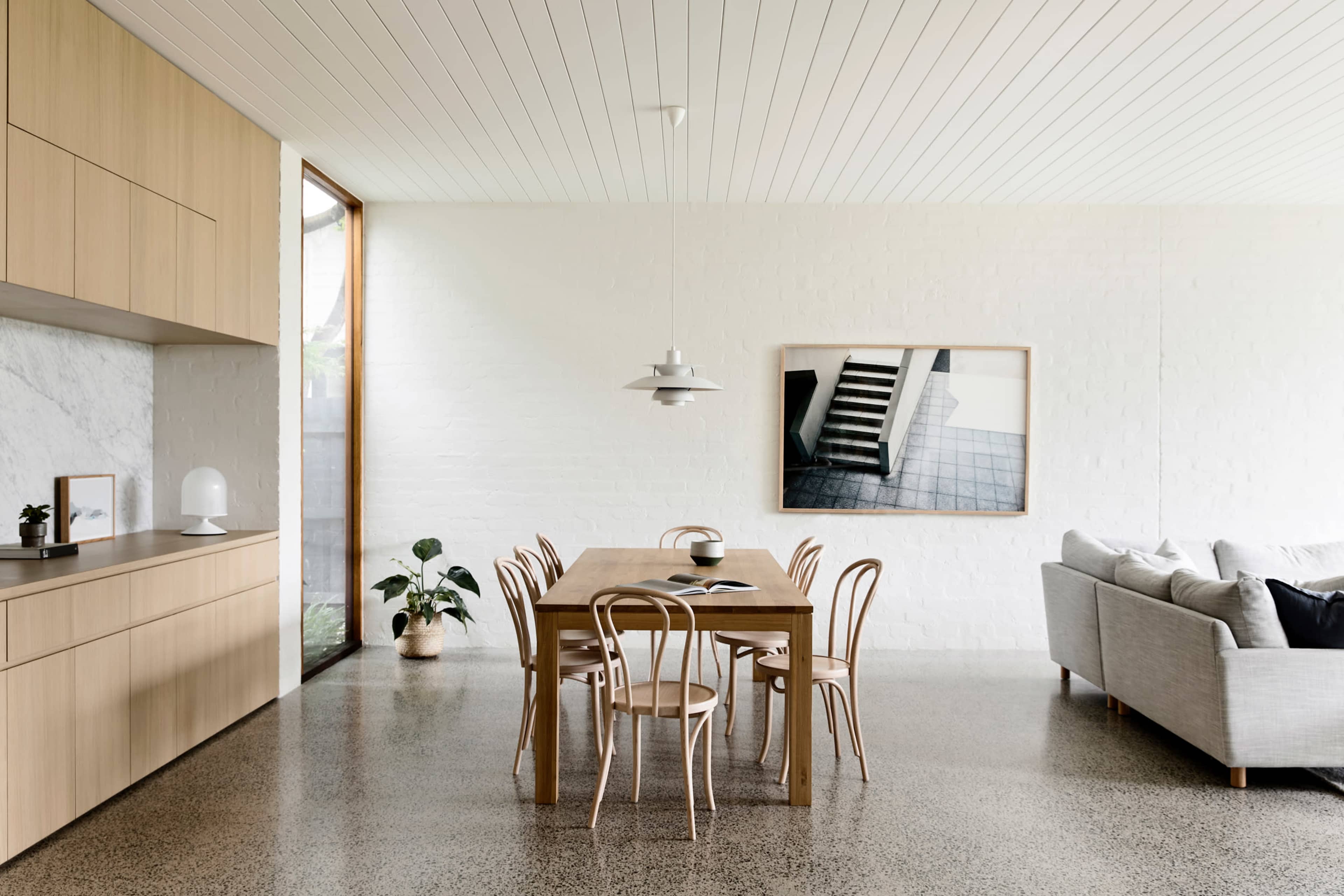 A modern dining area featuring a wooden table surrounded by chairs, large windows, a gray sofa, and a minimalist décor.