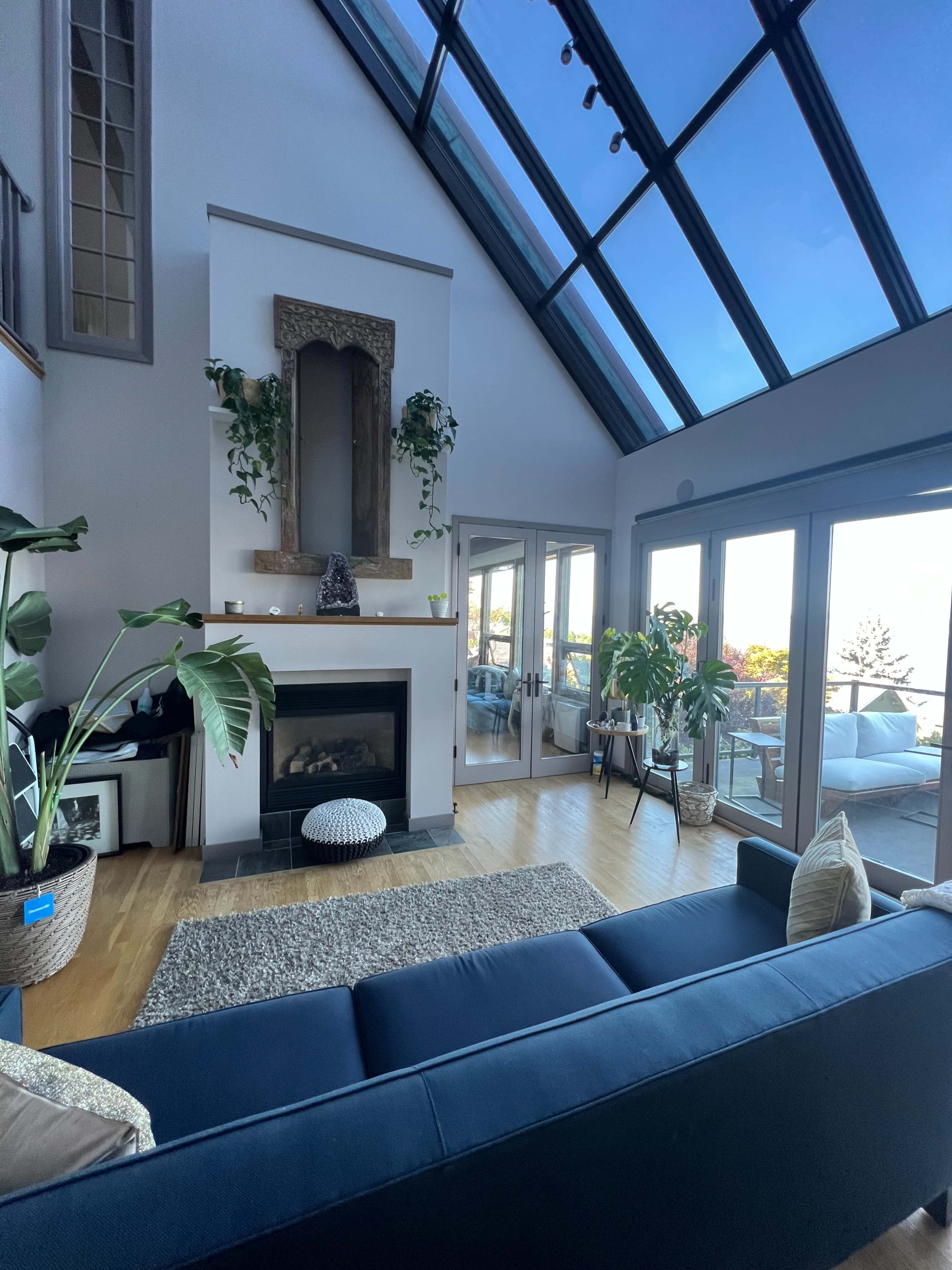 A bright living room features a large windowed wall, a fireplace, and plants around a dark sofa.