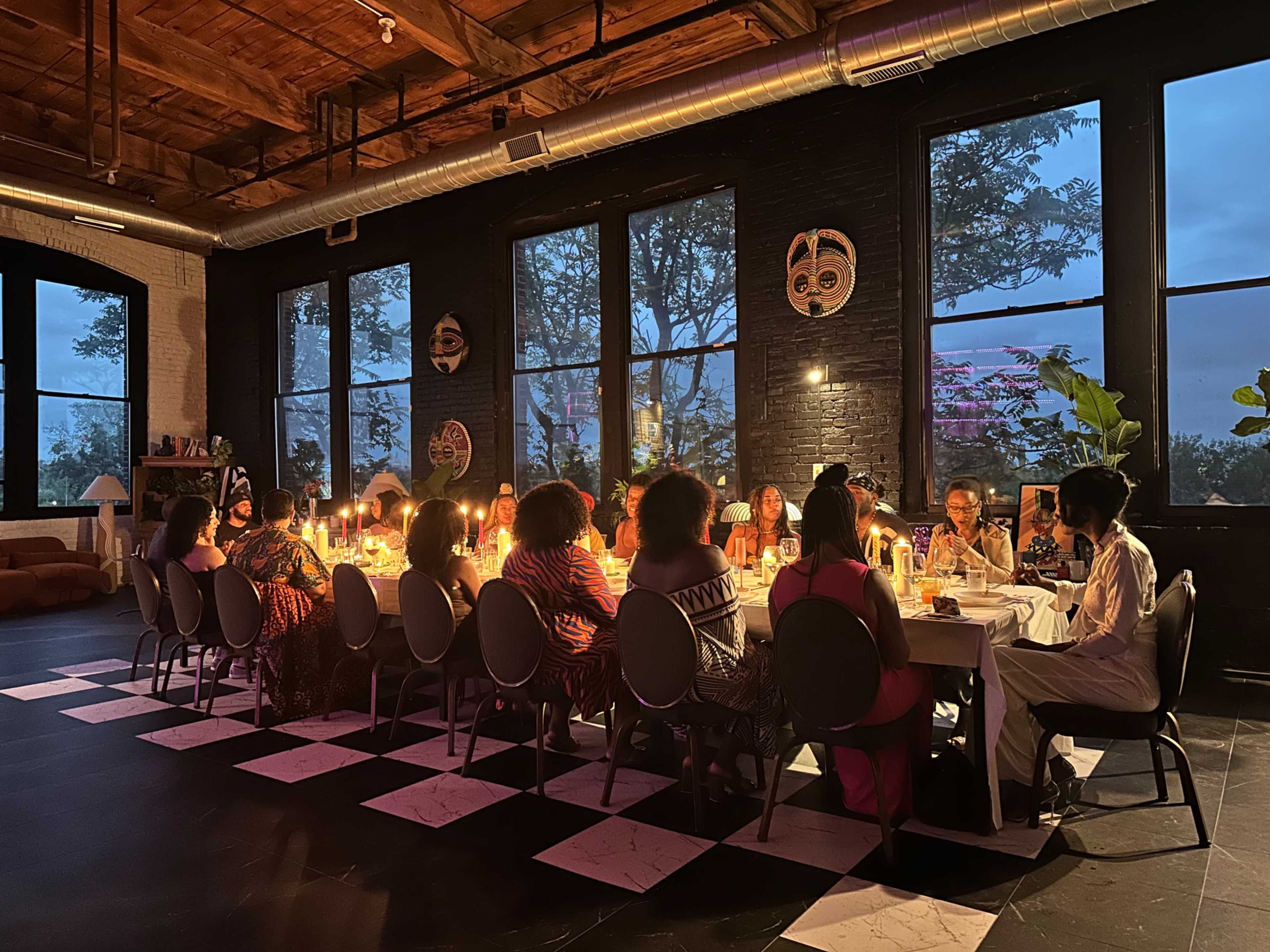 A long table set for a dinner gathering is surrounded by guests under dim lighting, with large windows displaying an evening sky.